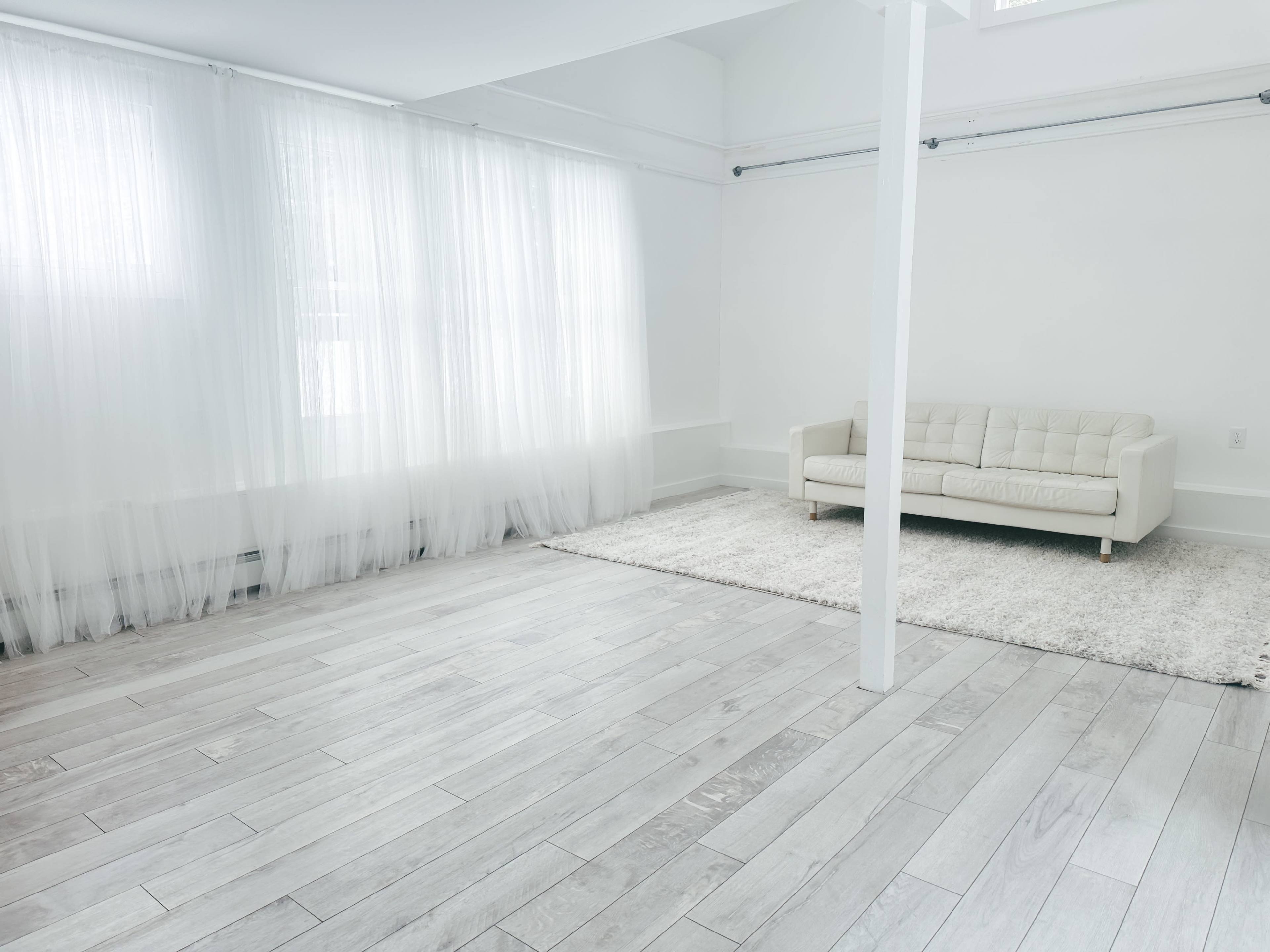 A bright, minimalist room with white walls, light-colored wooden flooring, a plush area rug, and a modern sofa near sheer curtains.
