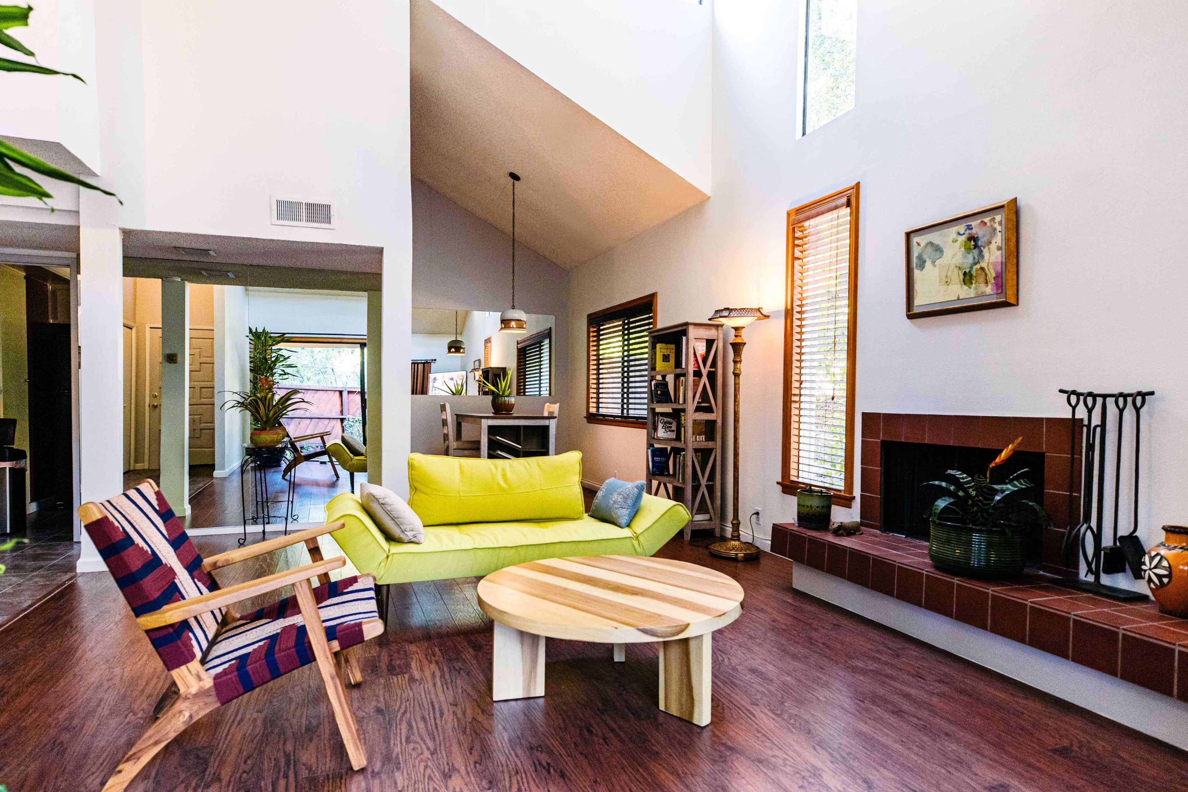 A modern living room features a yellow couch, a round wooden coffee table, and large windows with natural light.