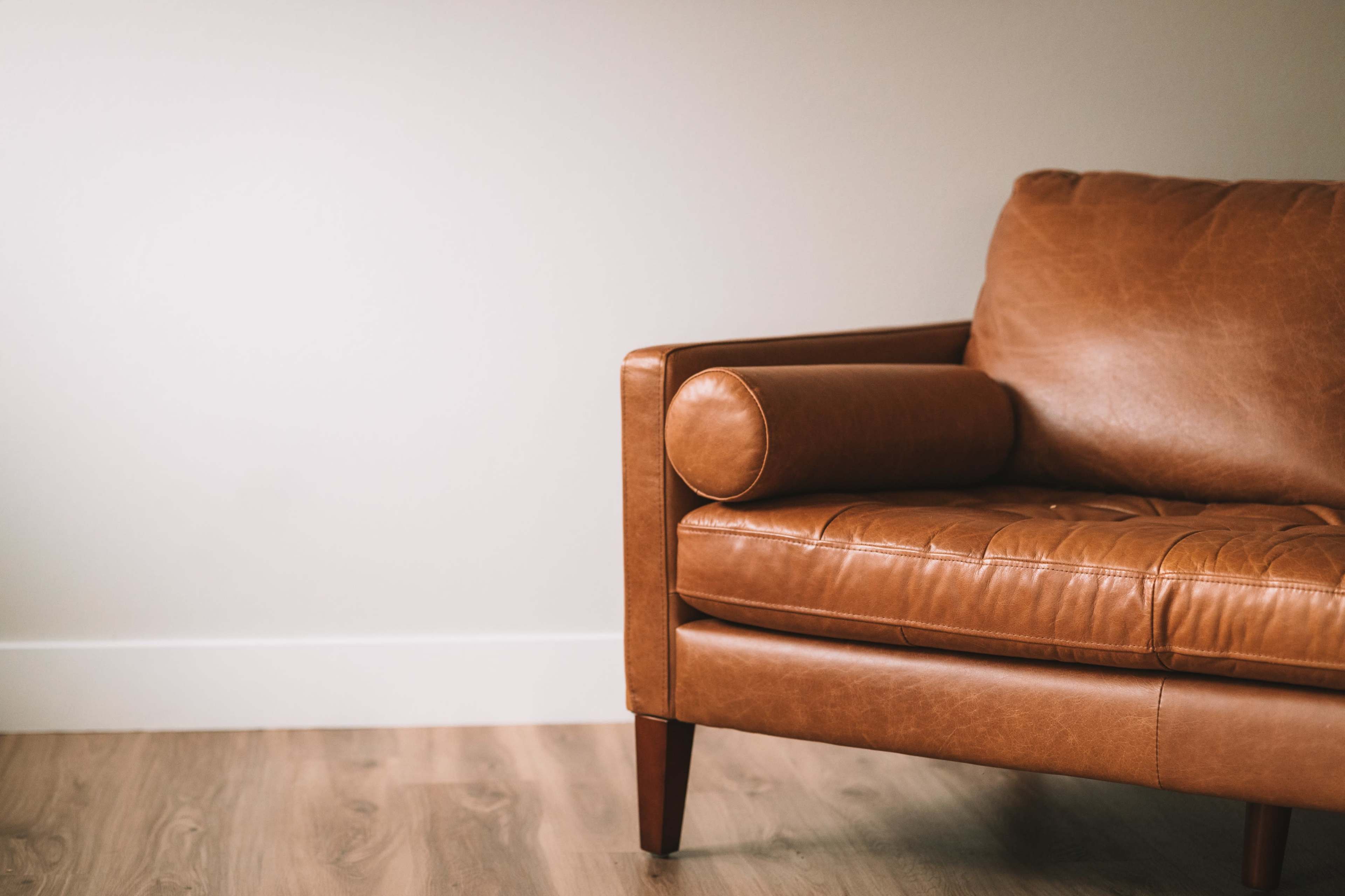 A brown leather couch with a round bolster cushion is positioned against a light-colored wall.