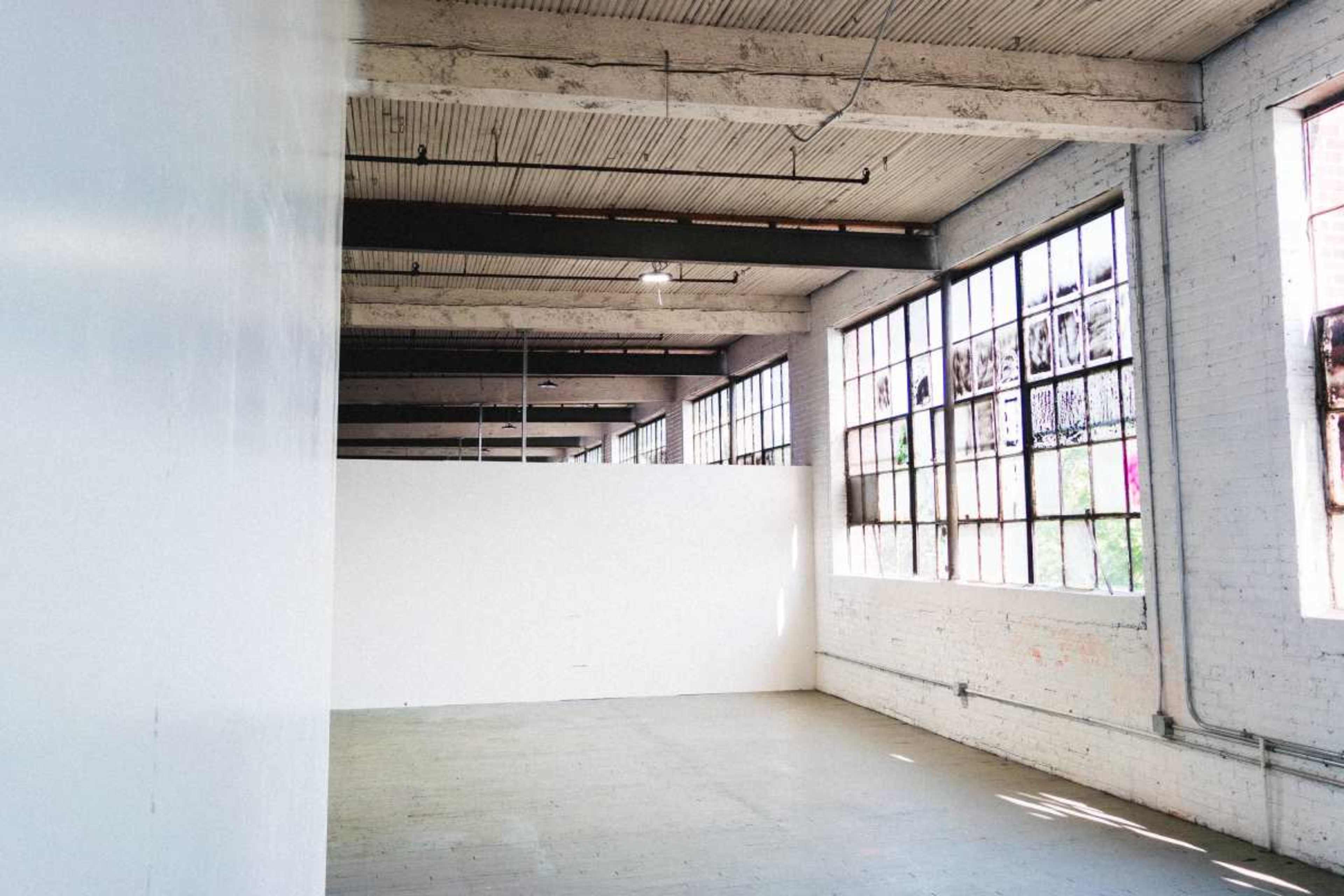 The image shows a spacious, industrial-style room with large windows and a blank wall dividing the space.