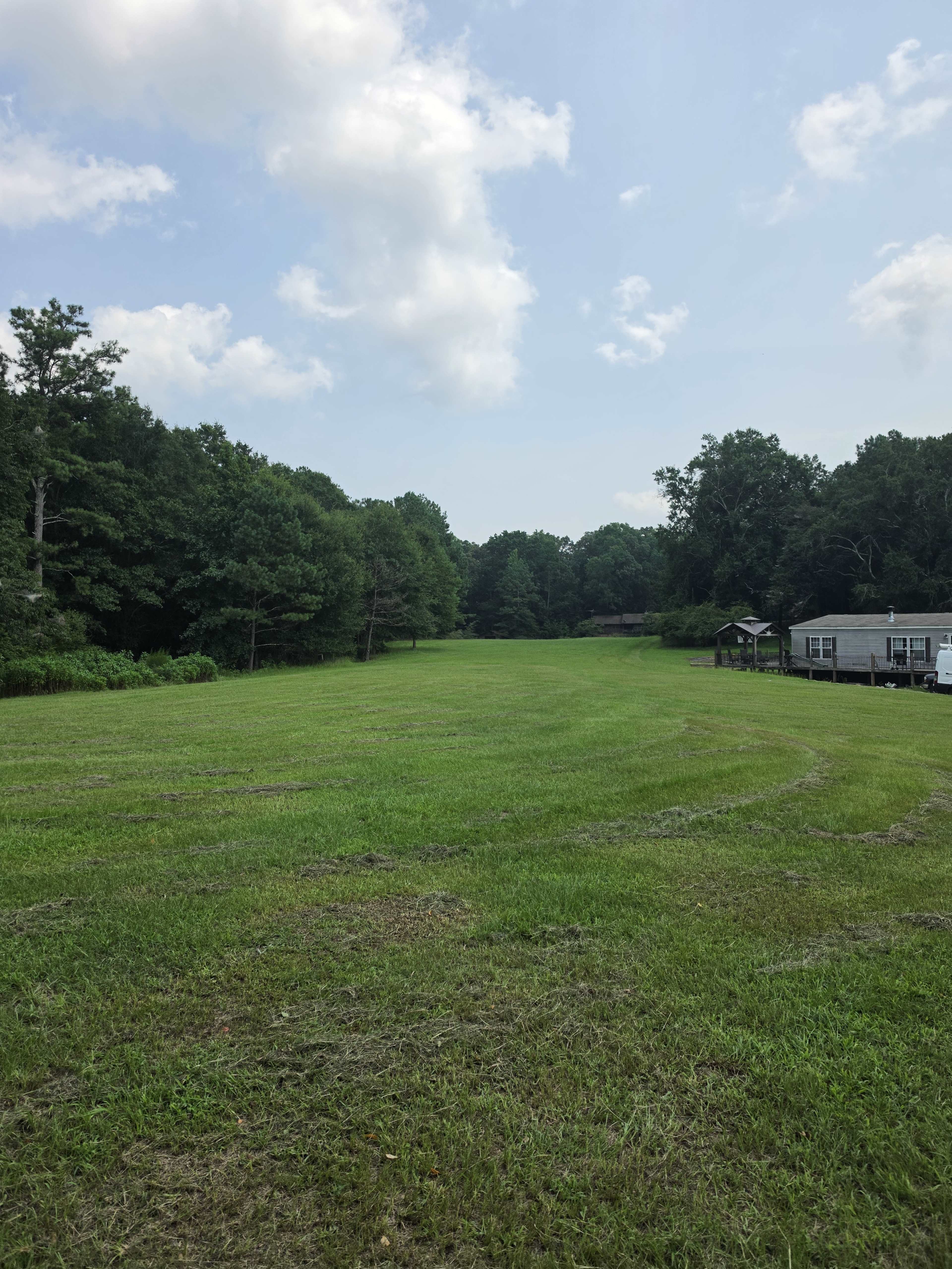 A wide, grassy field stretches between clusters of trees under a partly cloudy sky, with a building visible in the distance.