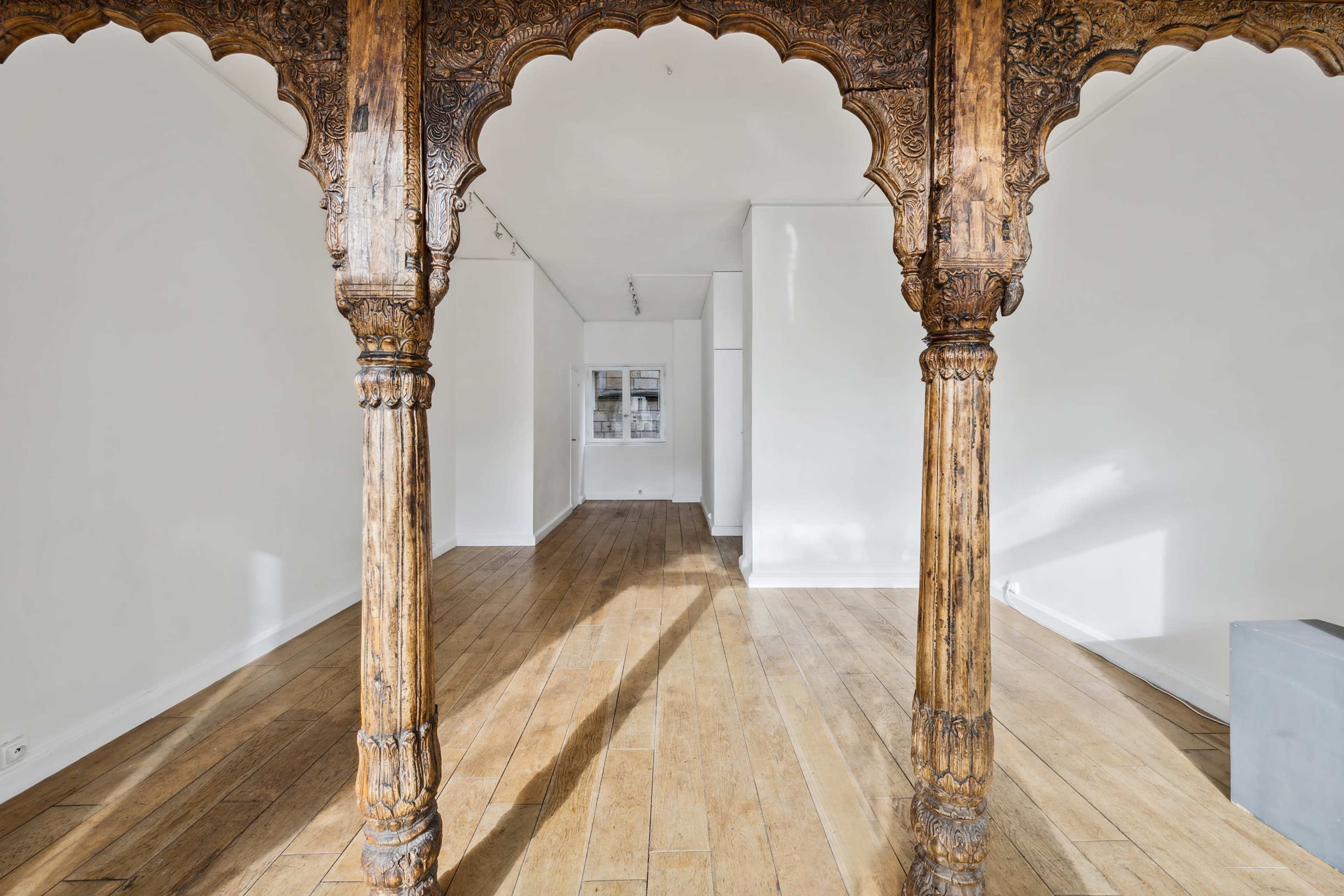 The image shows a spacious room with polished wooden floors and intricately carved wooden columns framing the entrance.