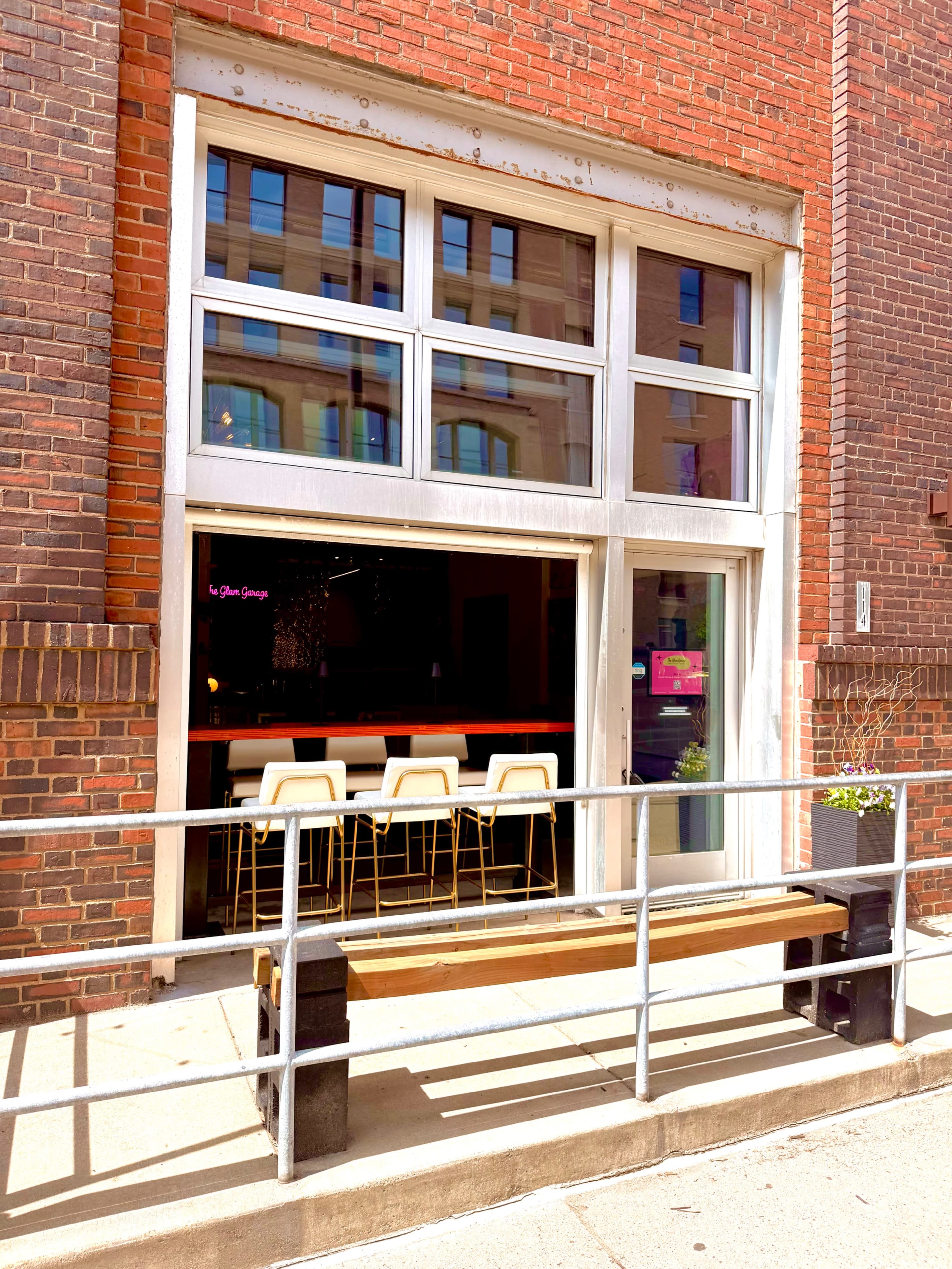 The image shows a storefront with large glass windows, featuring a row of white bar stools and a wooden bench in front.