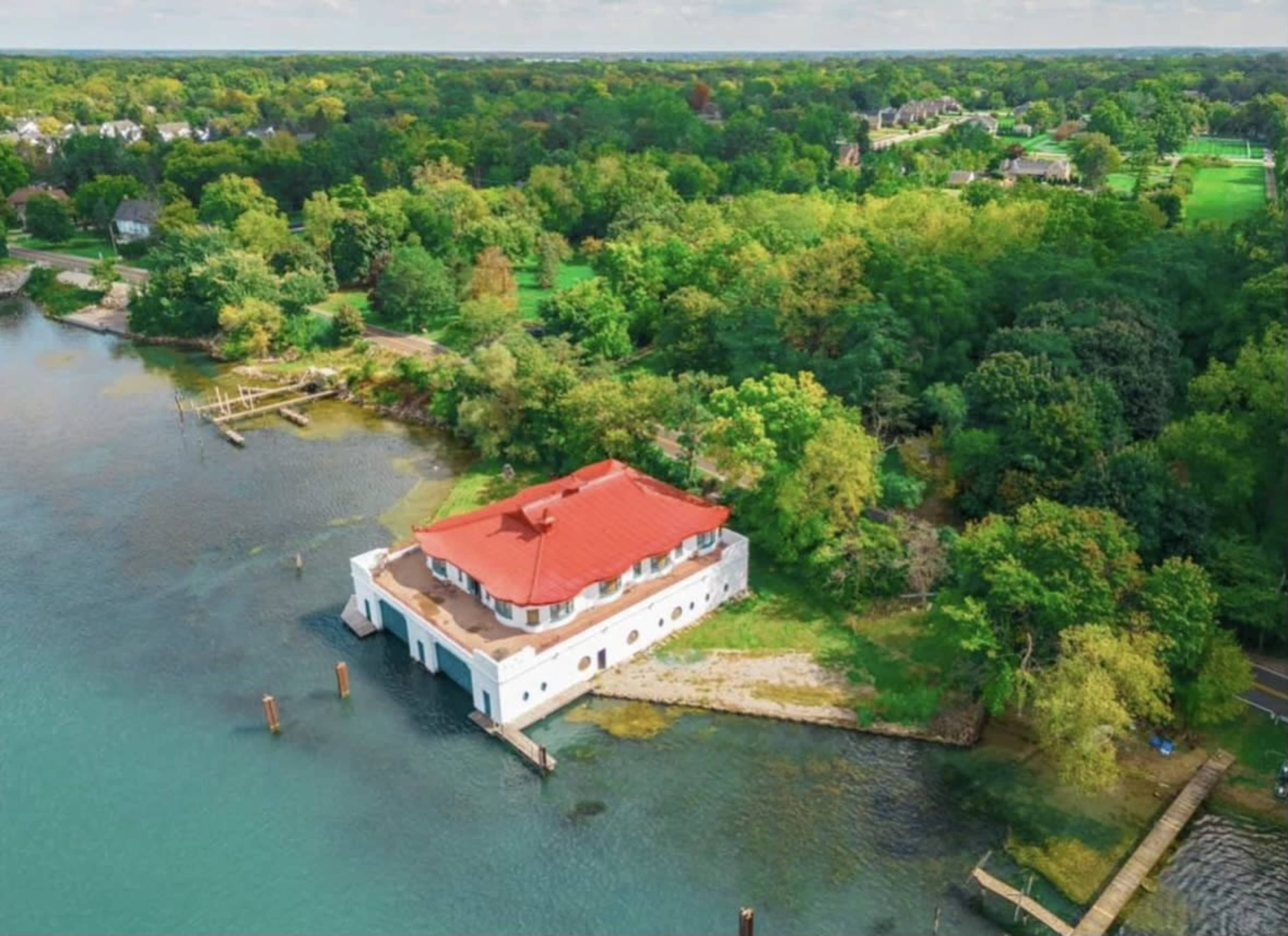 A large, white building with a red roof sits on the edge of a body of water, surrounded by lush greenery and a few scattered structures in the background.