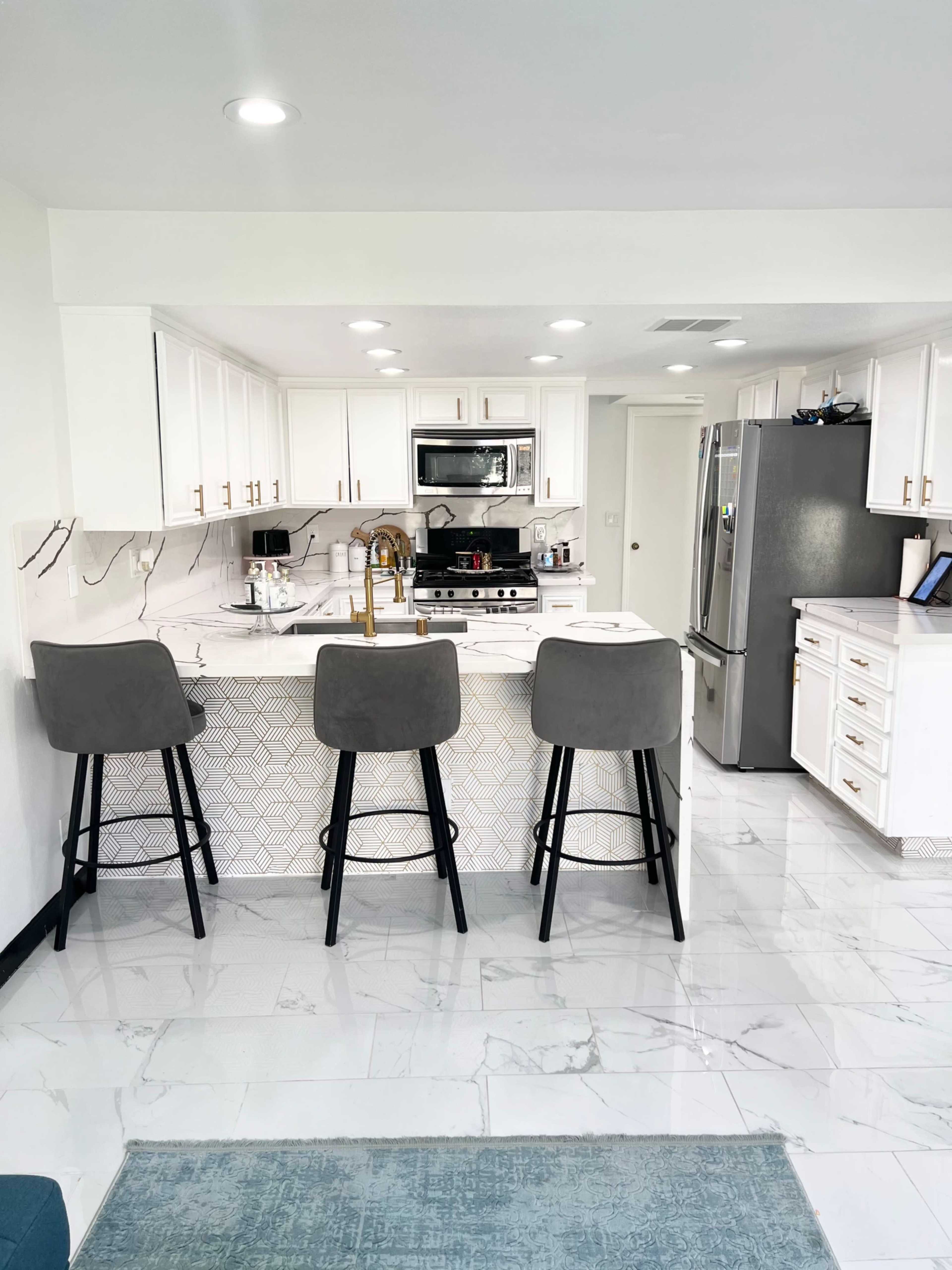 A modern kitchen features a central island with three high stools, stainless steel appliances, and white cabinetry accented by gold hardware.