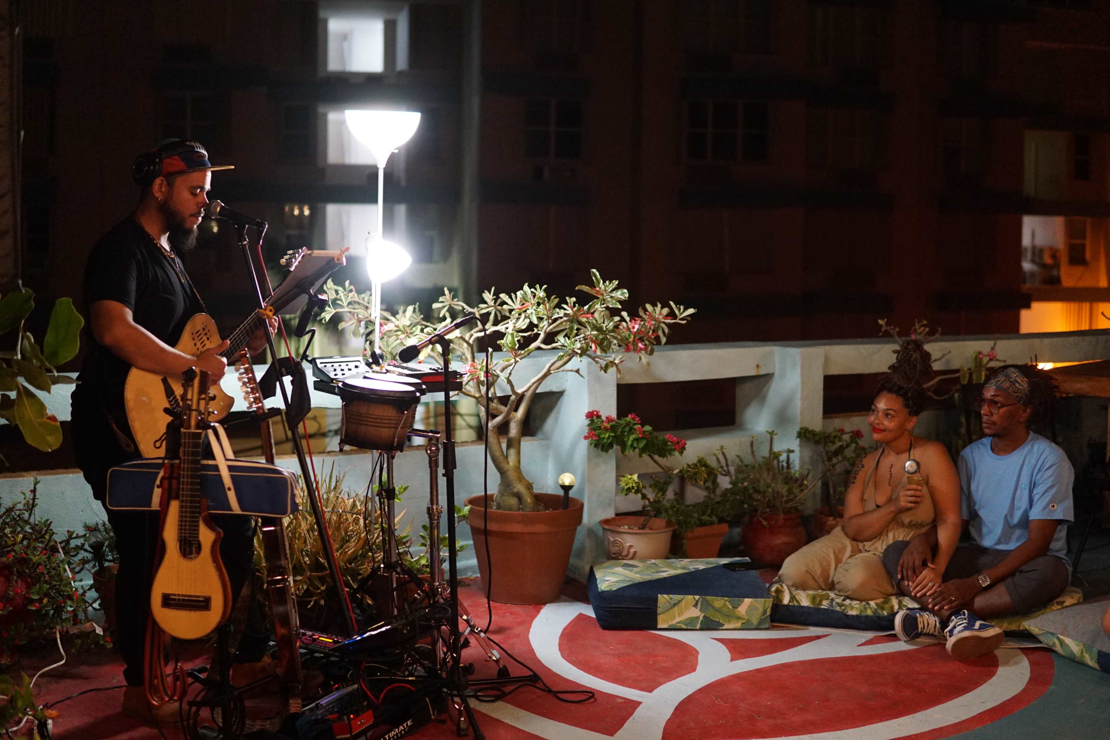 A musician performs with a guitar and percussion instruments on a rooftop while two audience members sit nearby, surrounded by potted plants and decorative cushions.
