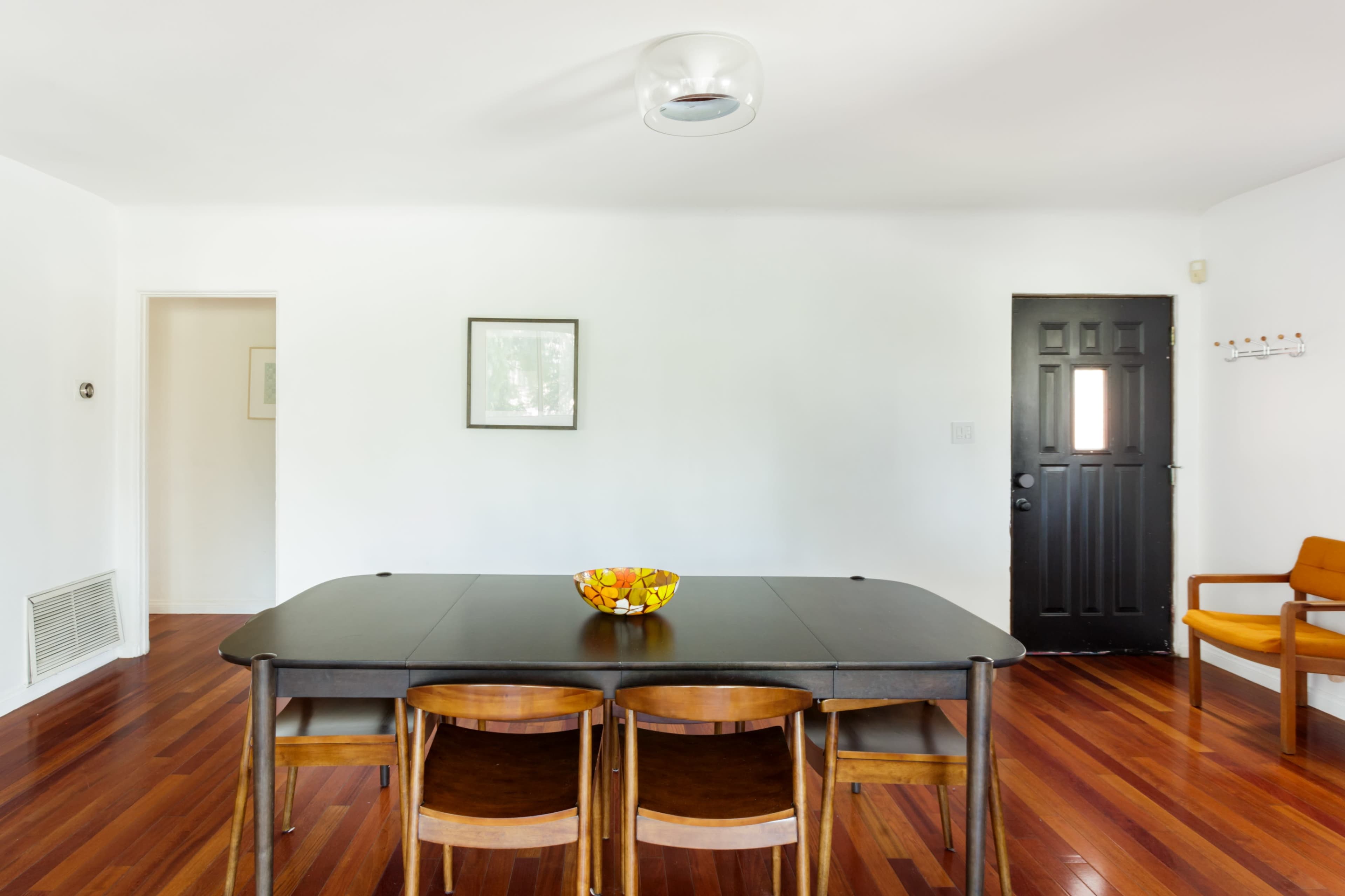 A modern dining area features a dark wooden table surrounded by four chairs, with a bowl of fruit at the center and a black door leading outside.