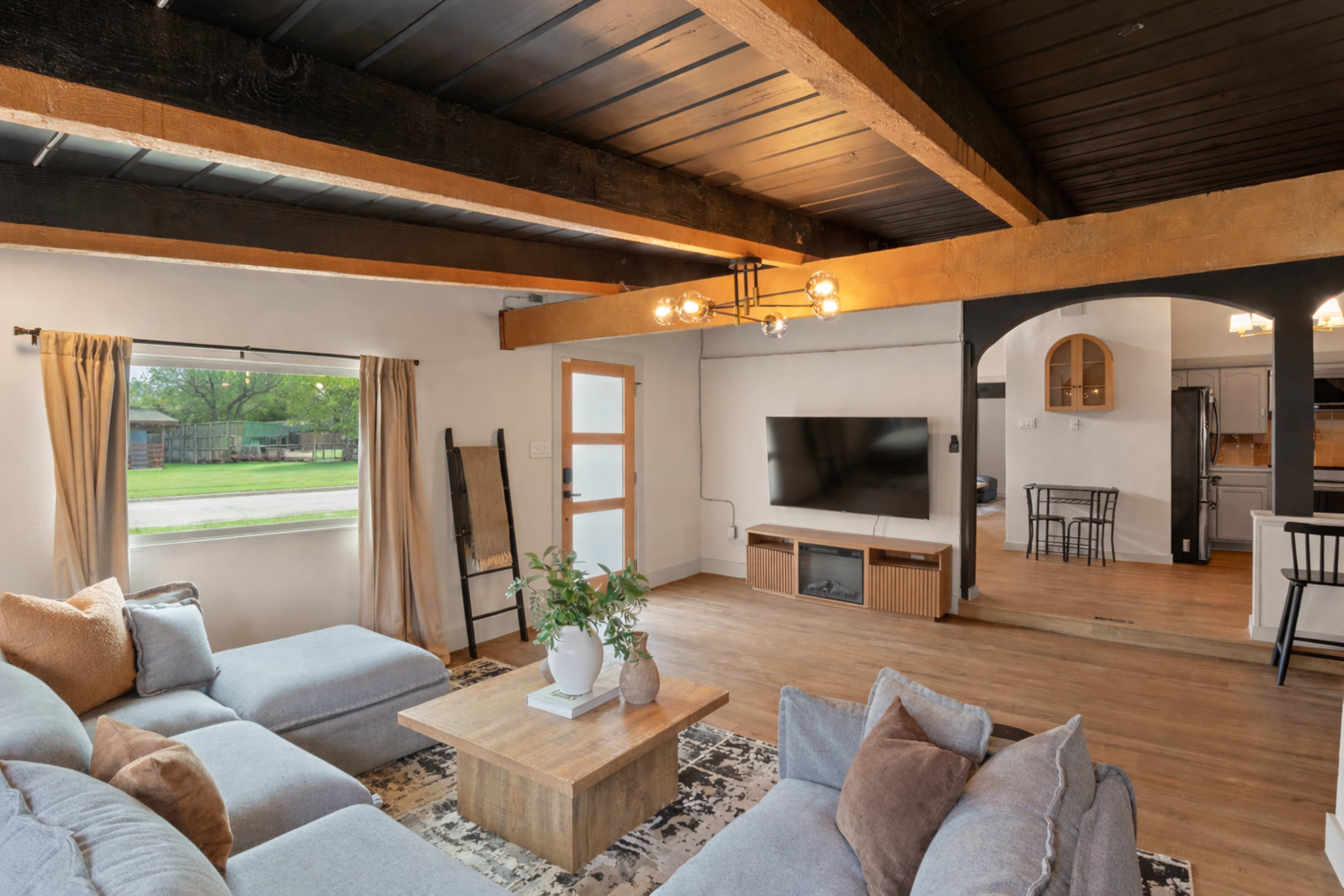 The image shows a cozy living room with a light gray sectional sofa, a wooden coffee table, and a wall-mounted television, framed by wooden beams above and large windows allowing natural light.