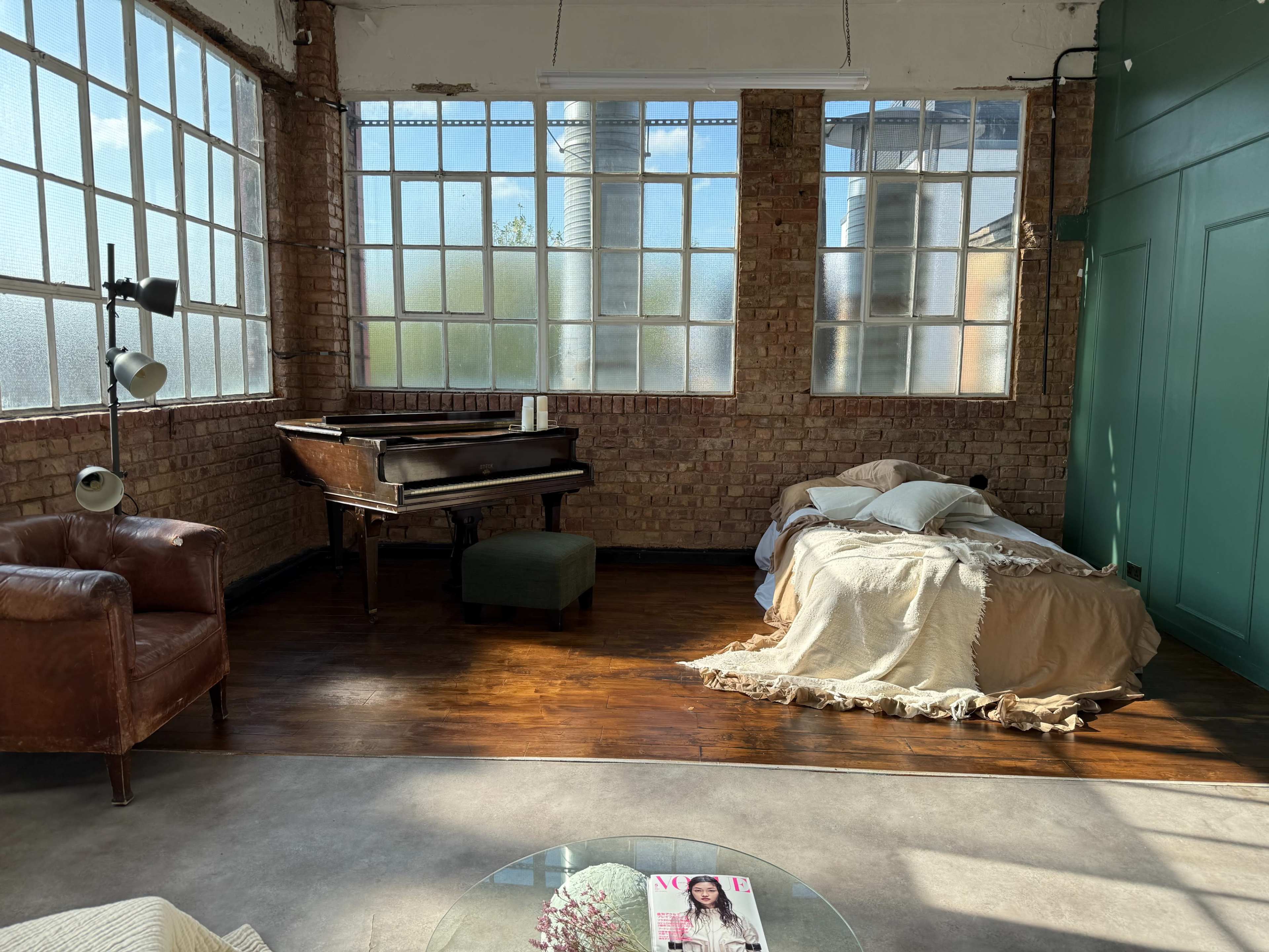 The image shows a bright, spacious room with large windows, featuring a piano, a leather armchair, and a bed draped in blankets.