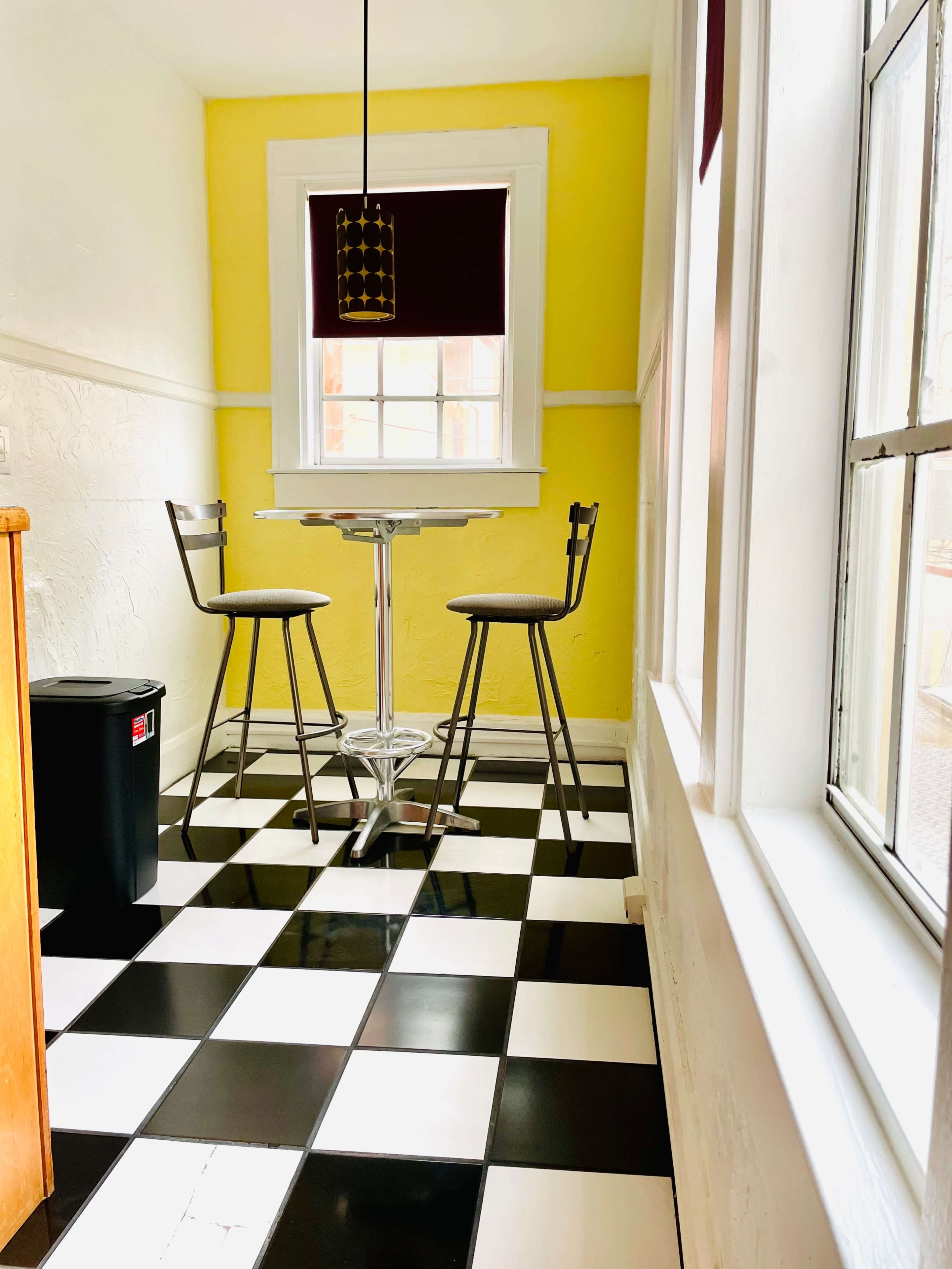 A small kitchen nook features a round table and two high stools against a bright yellow wall, with black and white checkered flooring.