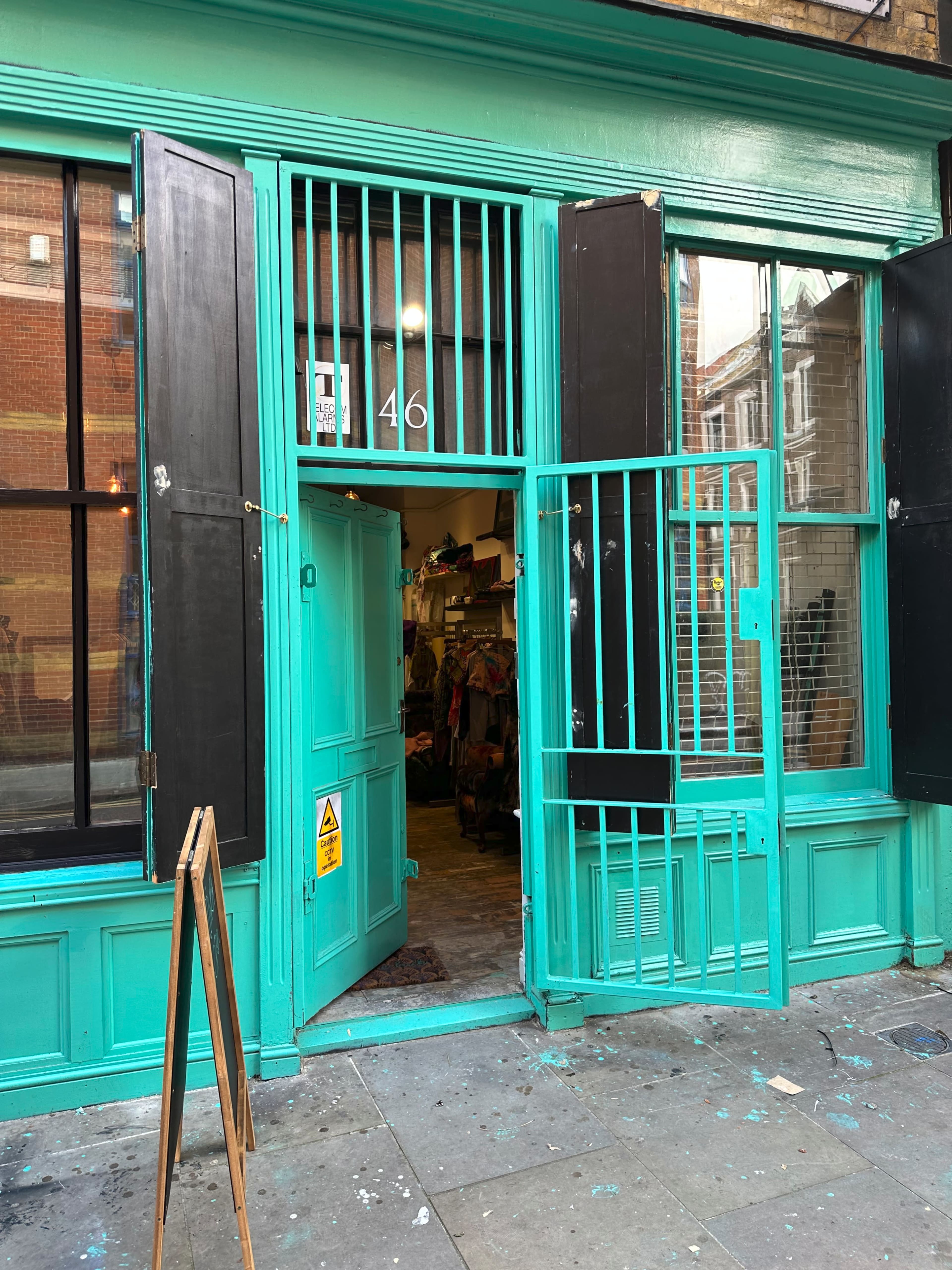 A turquoise-painted shopfront with an open door, featuring black shutters and a small sign outside.