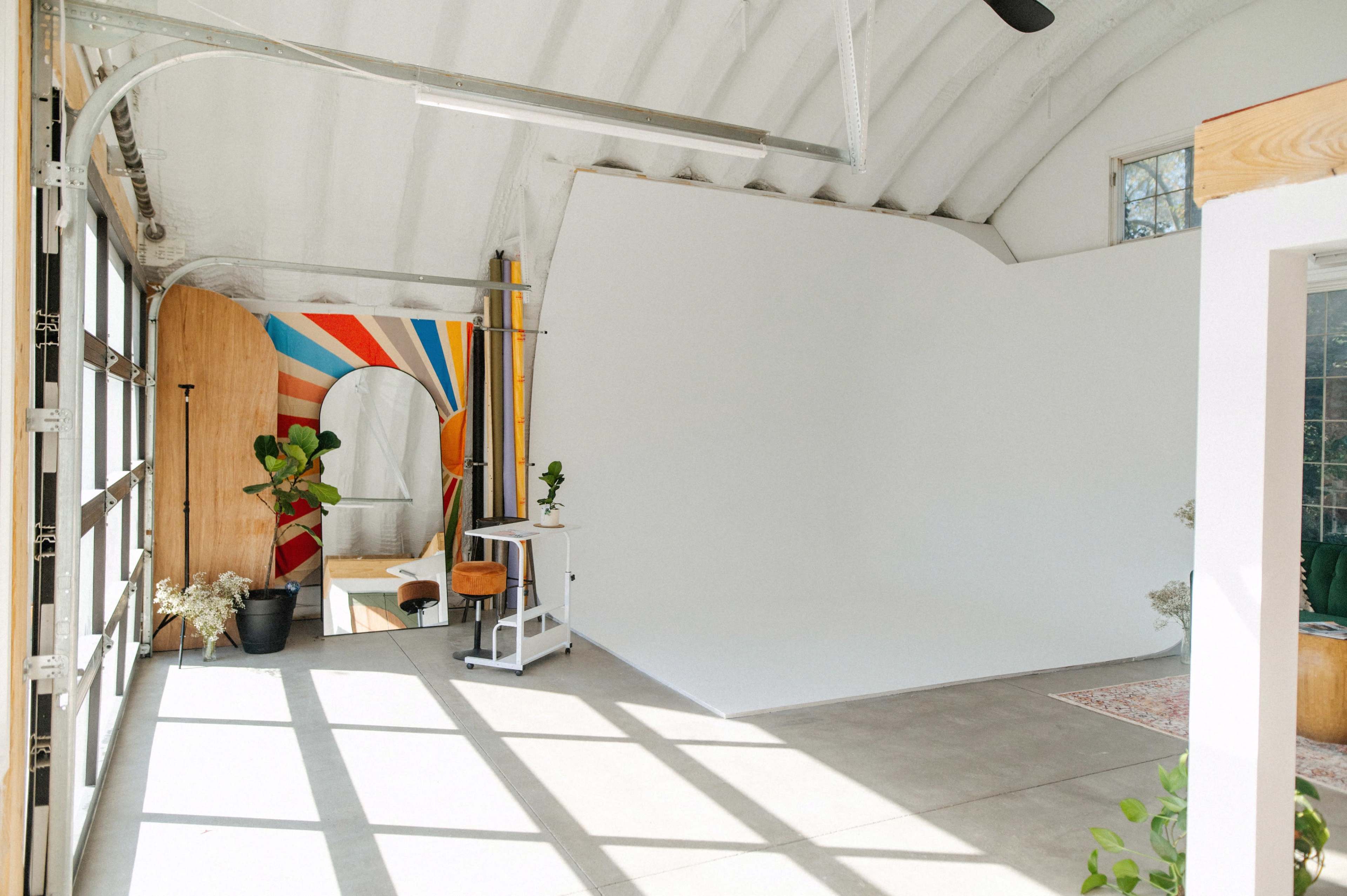 An indoor studio space featuring a large white backdrop, colorful wall art, and minimal furniture including a stool and a small table.