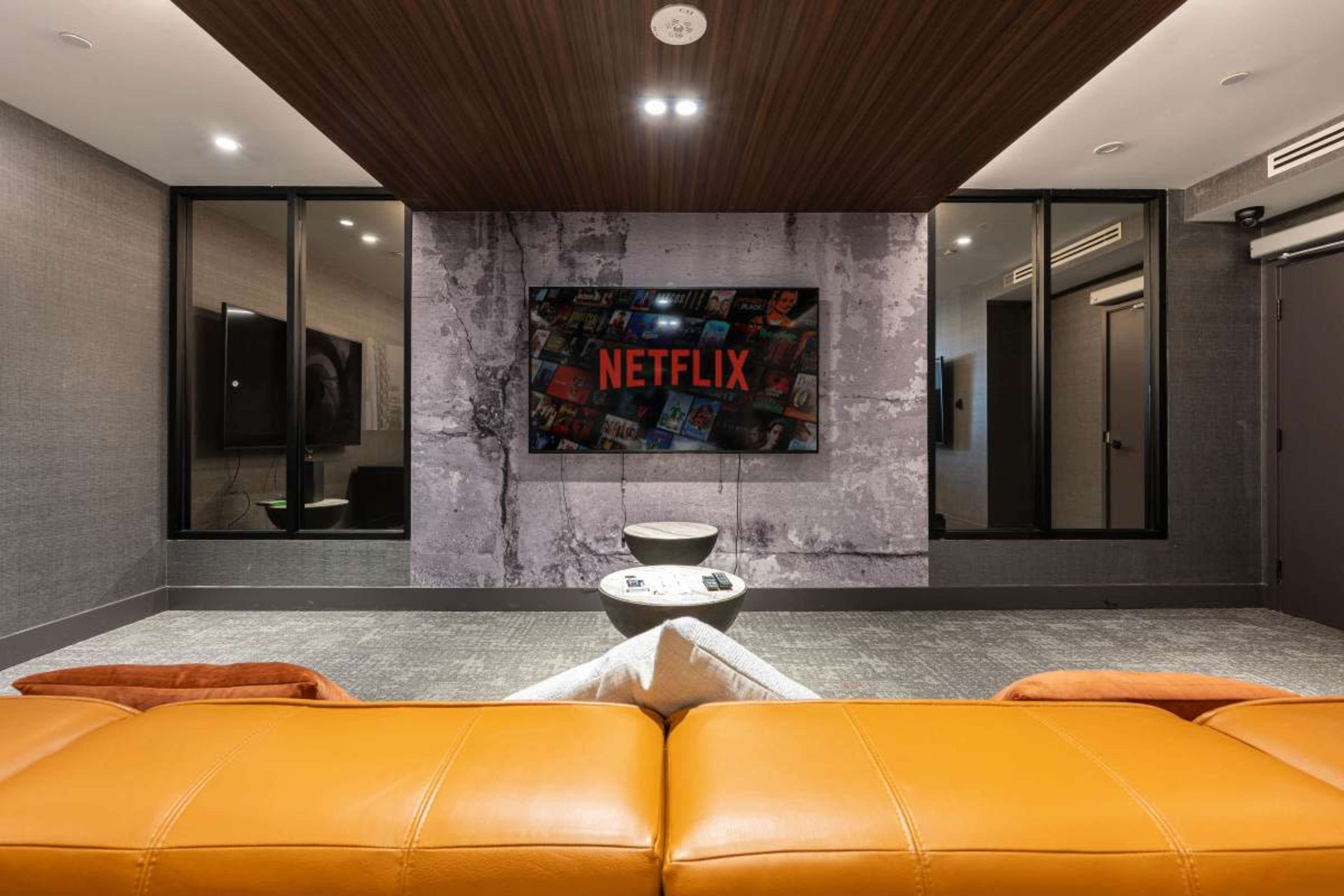 The image shows a modern lounge area featuring a large television displaying the Netflix logo, with a sleek orange couch and decorative wall.