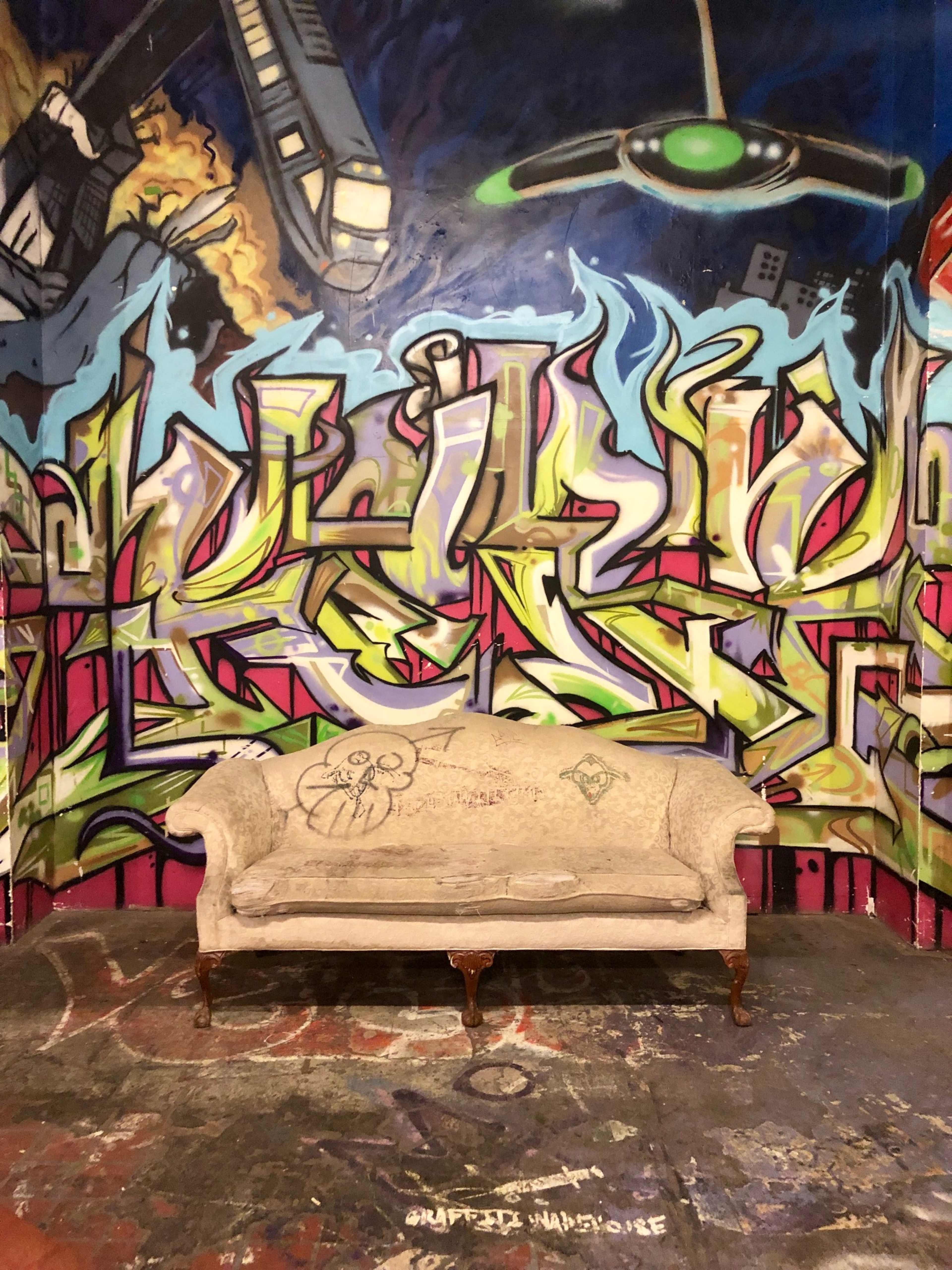 A vintage couch positioned in front of a vibrant, graffiti-covered wall in an urban setting.