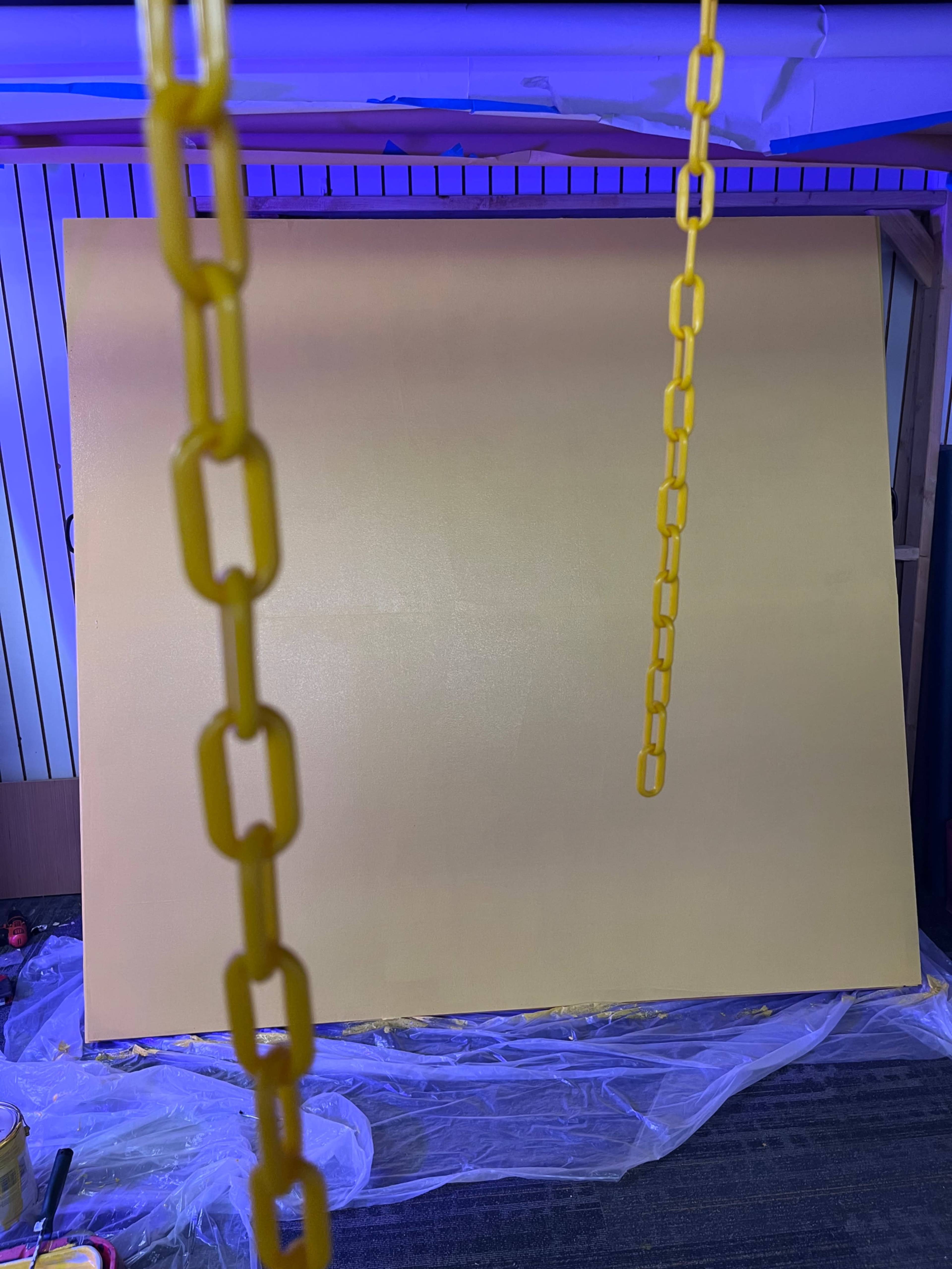 A large yellow canvas is suspended in front of a backdrop, with yellow chains hanging from above and paint supplies visible below.