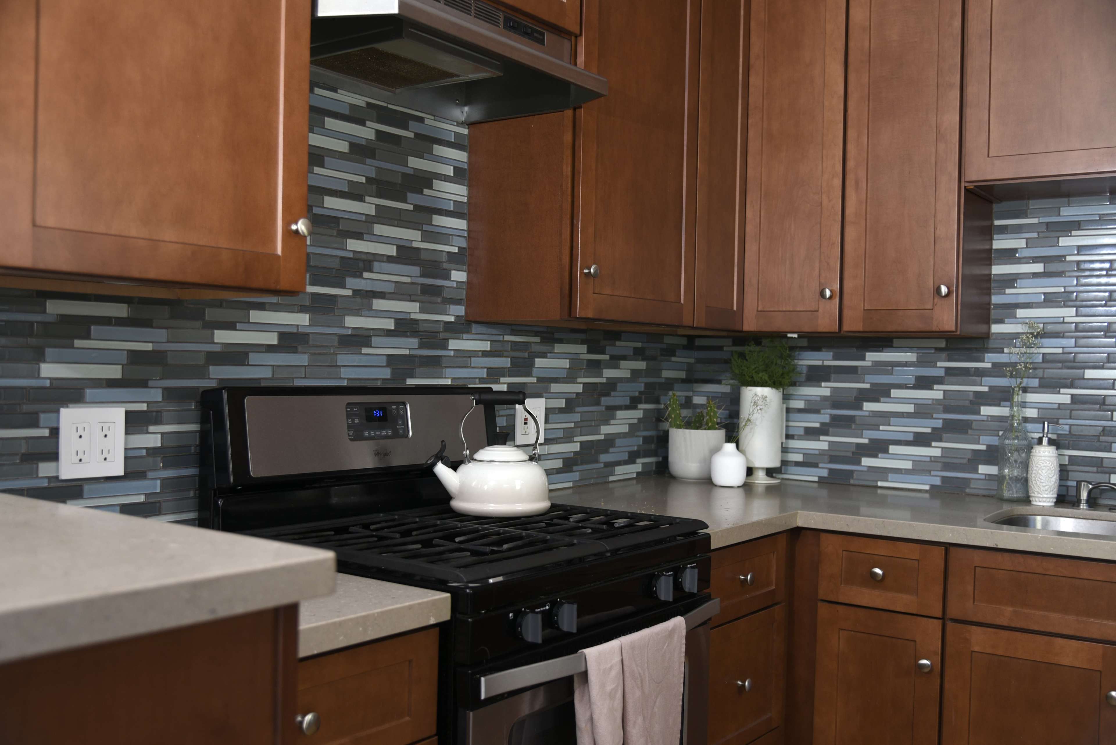 A kitchen with wooden cabinets, a gas stove with a kettle, and a tiled backsplash featuring varying shades of blue and gray.