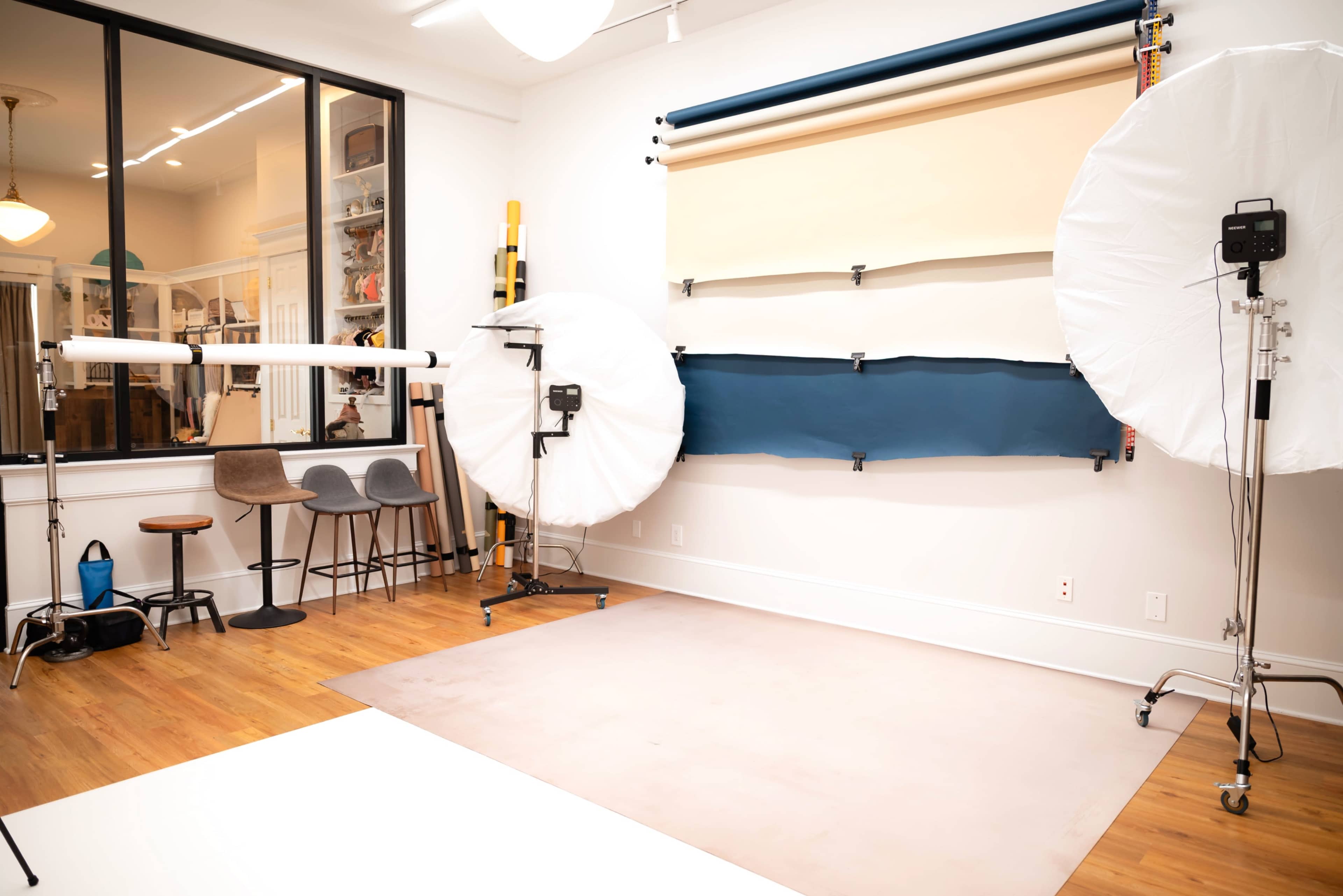 The image shows a well-lit photography studio featuring backdrops, lighting equipment, and a seating area with three stools.