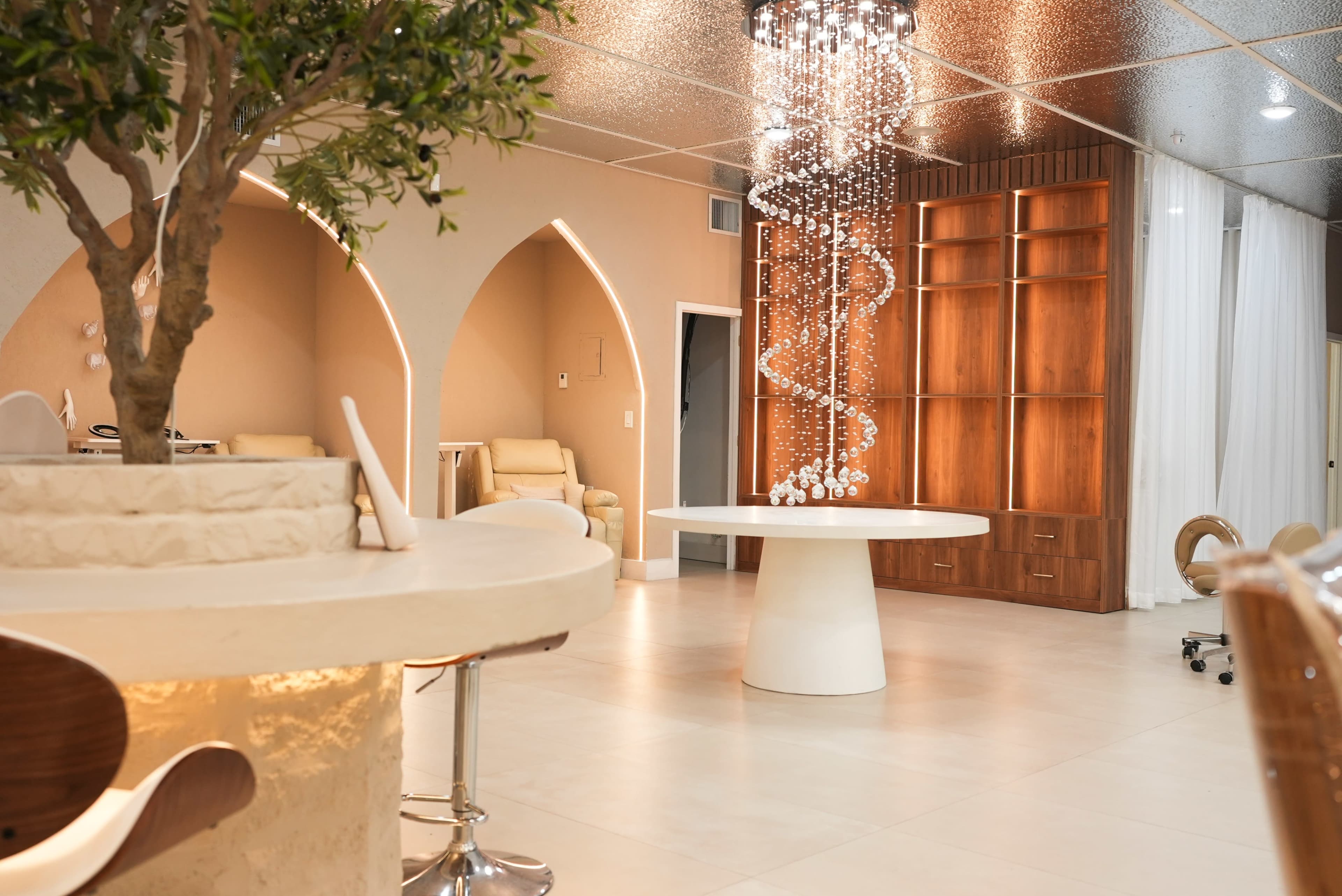 The image shows a modern interior space featuring a circular white table, a bar-style seating area, and a wooden shelving unit, all illuminated by decorative lighting.