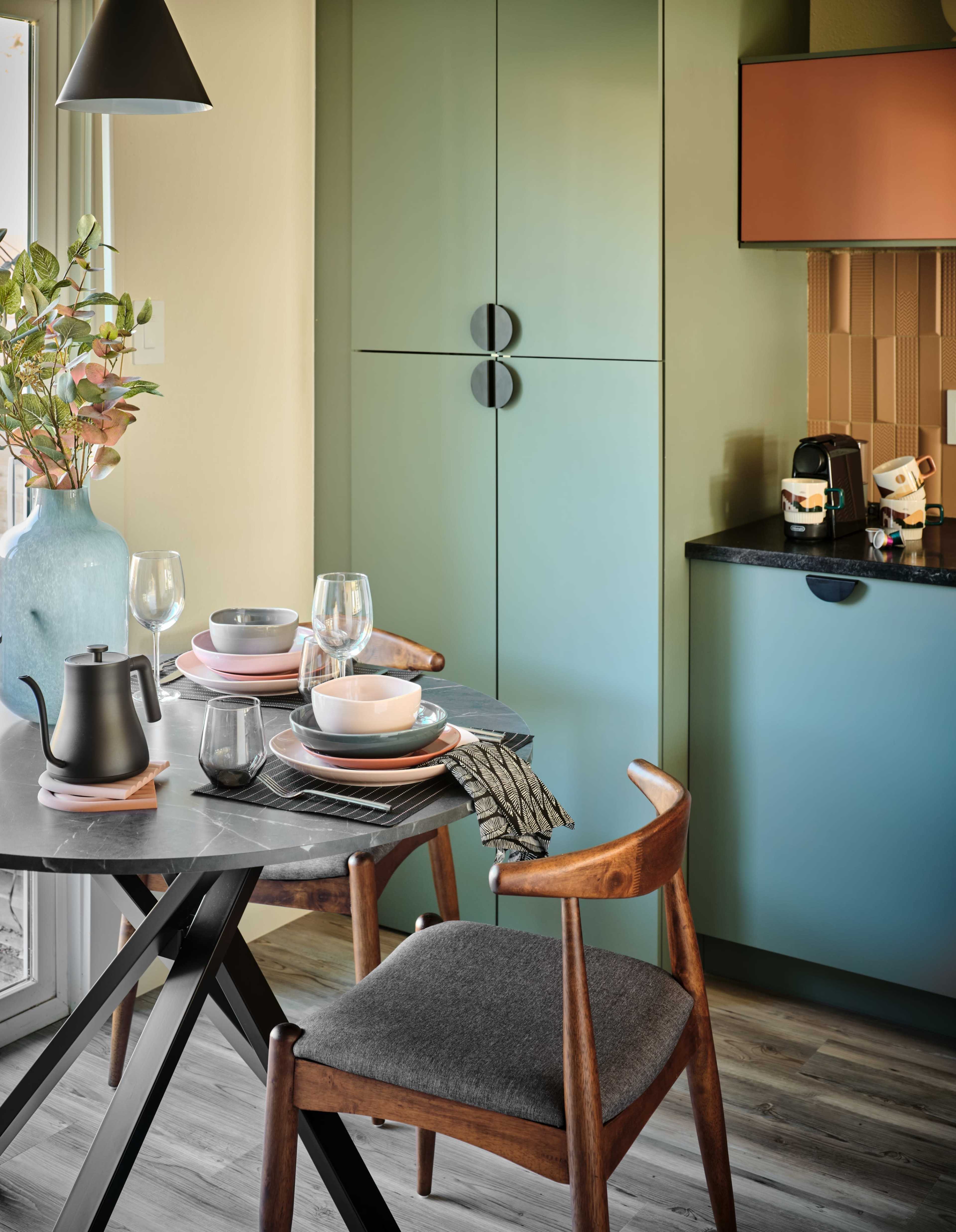 The image shows a modern kitchen area featuring a round black table set with dishes and glassware, complemented by a wooden chair, and colorful cabinets in the background.