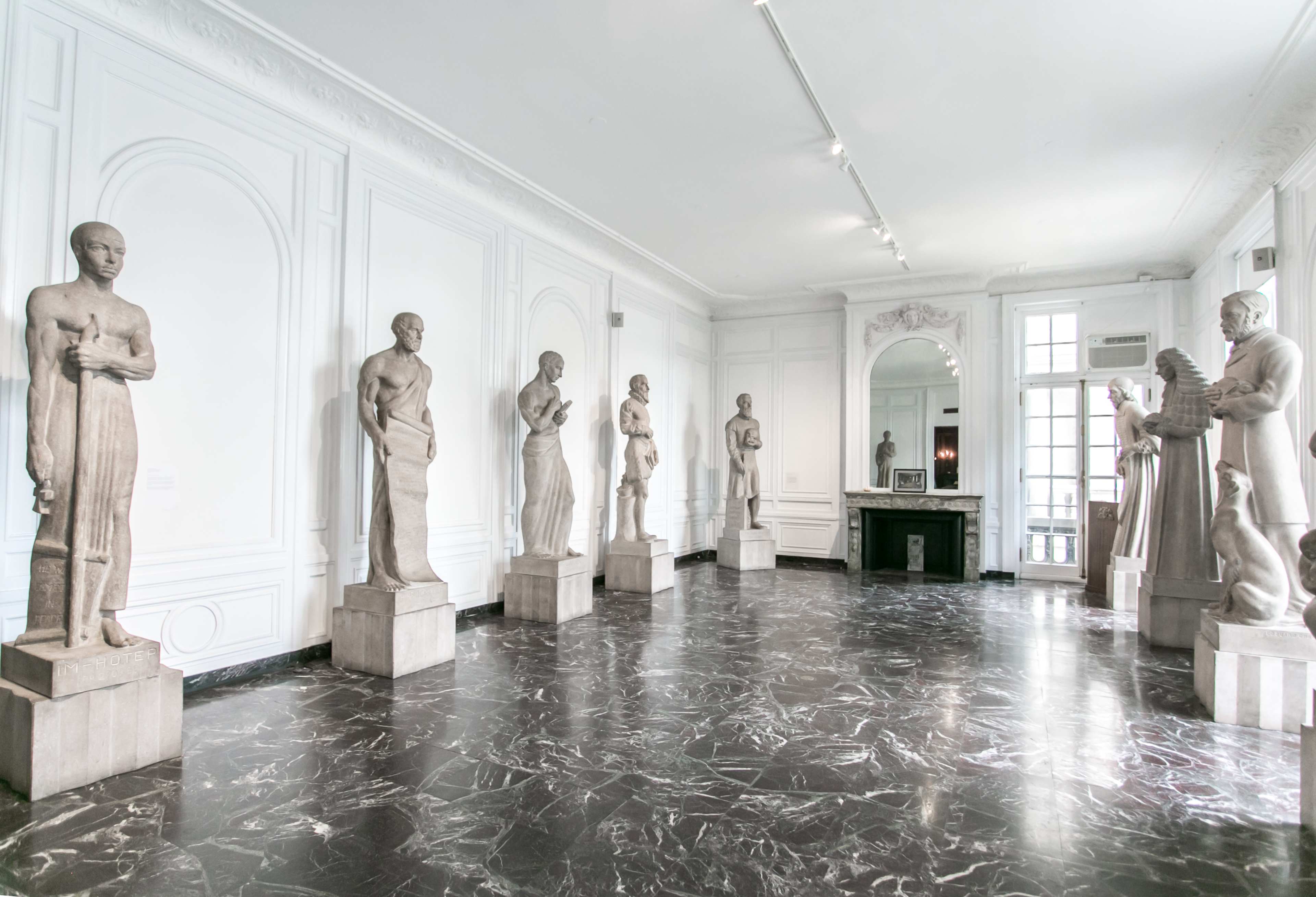 A gallery with several statues arranged on pedestals in a room featuring white walls and a dark marble floor.