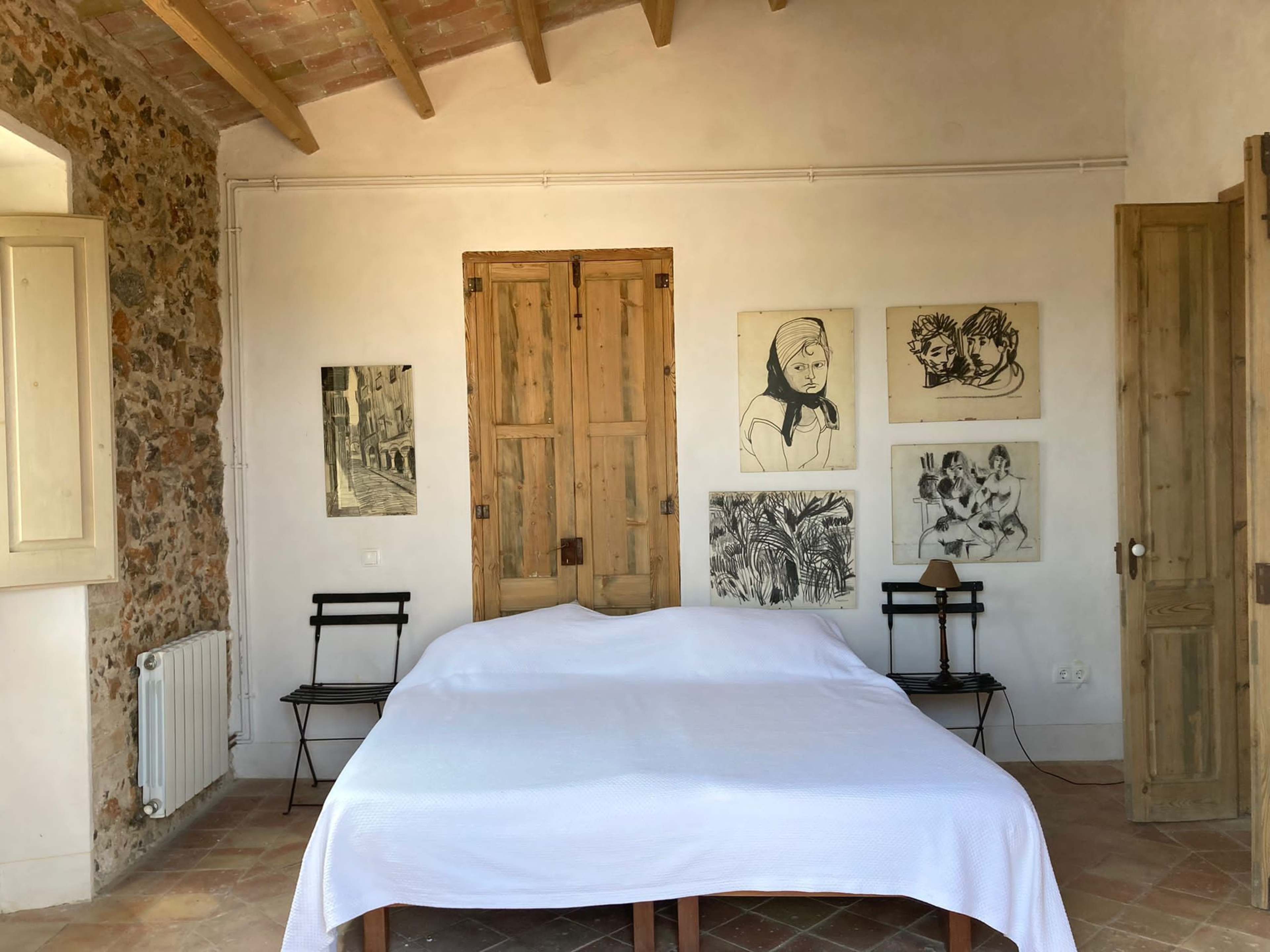 The scene features a simple bedroom with a wooden bed covered by a white sheet, flanked by paintings on the walls and wooden shutters, along with two foldable chairs.