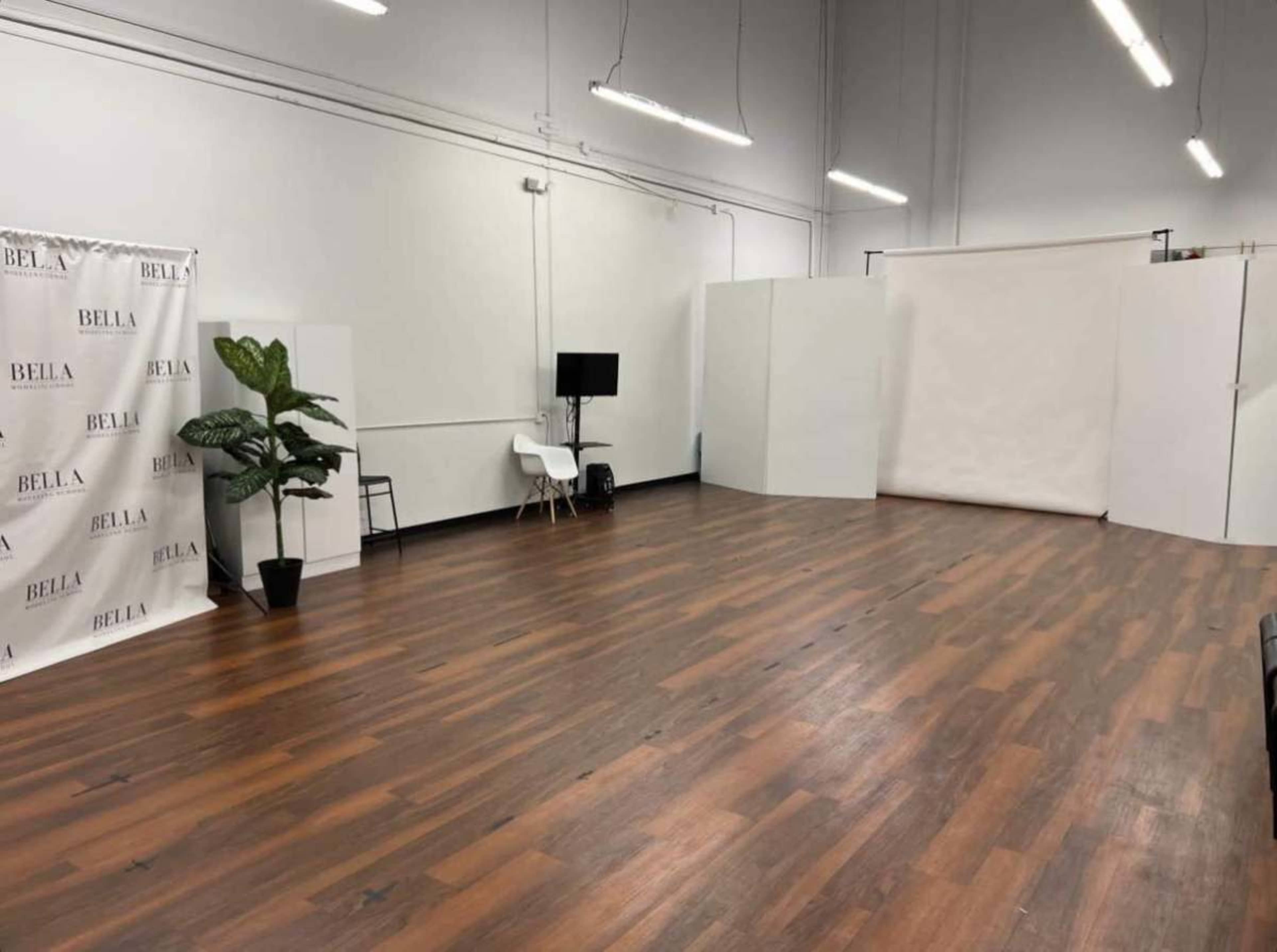 The image shows an empty studio space with hardwood floors, a backdrop featuring the name "BELLA," a potted plant, and minimal furniture.