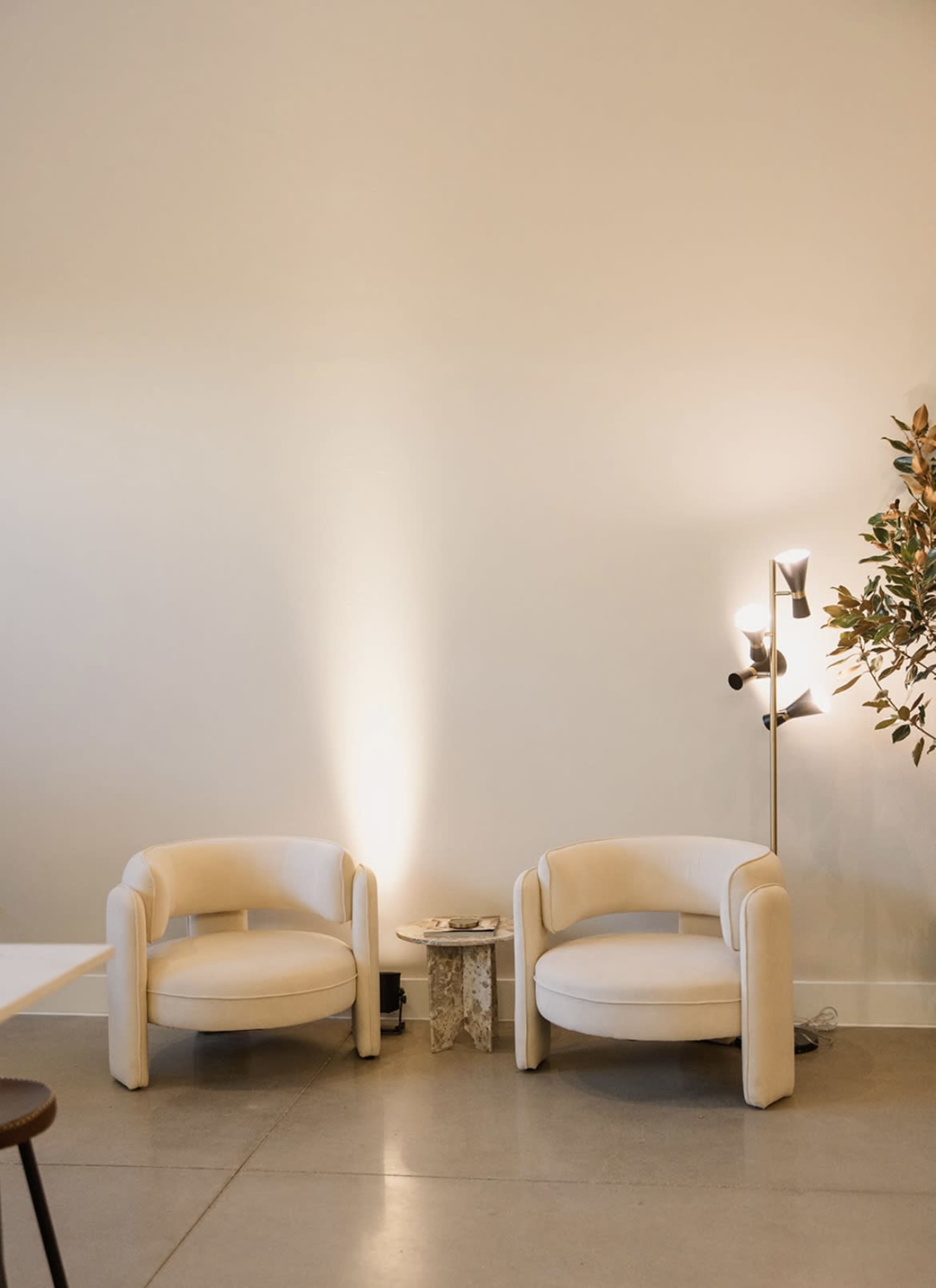 The image features two white, rounded armchairs positioned opposite each other, with a small table between them, against a blank wall illuminated by soft light.