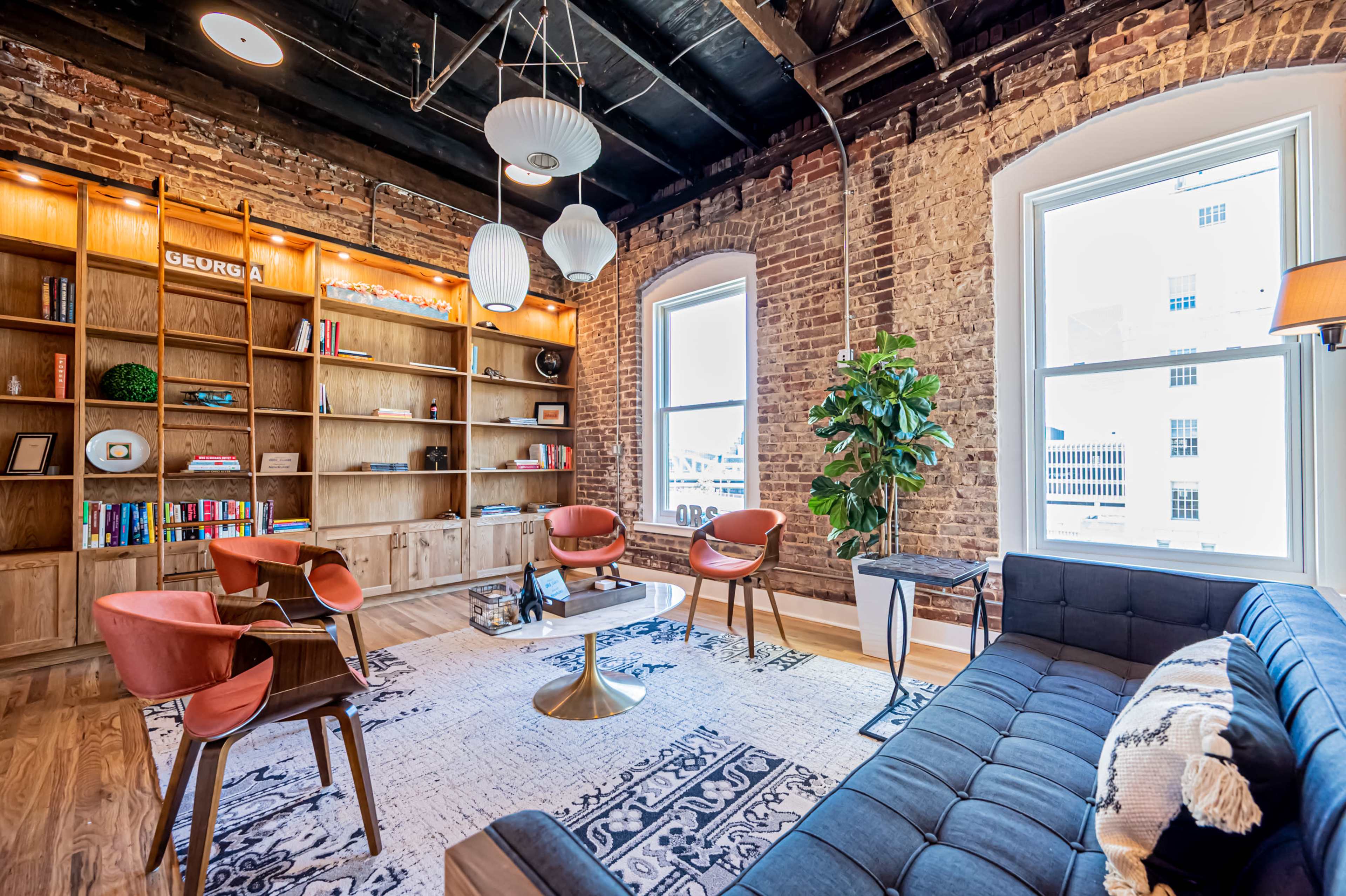 A modern lounge area featuring a sectional sofa, four orange chairs, a round coffee table, a bookshelf filled with books, and large windows allowing natural light to enter.