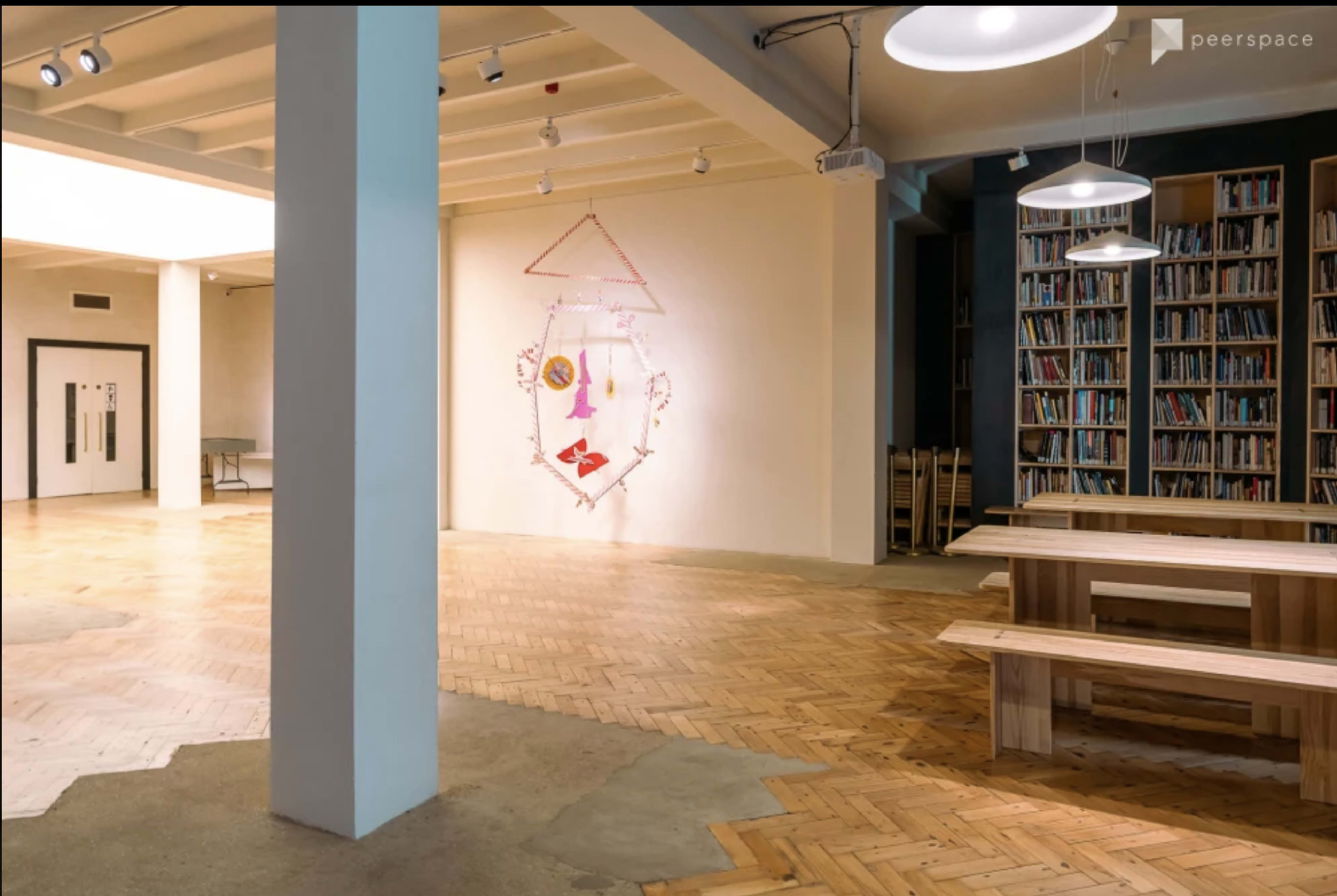 A spacious indoor area with wooden flooring, featuring a wall art installation, bookshelves, and a long wooden table with benches.