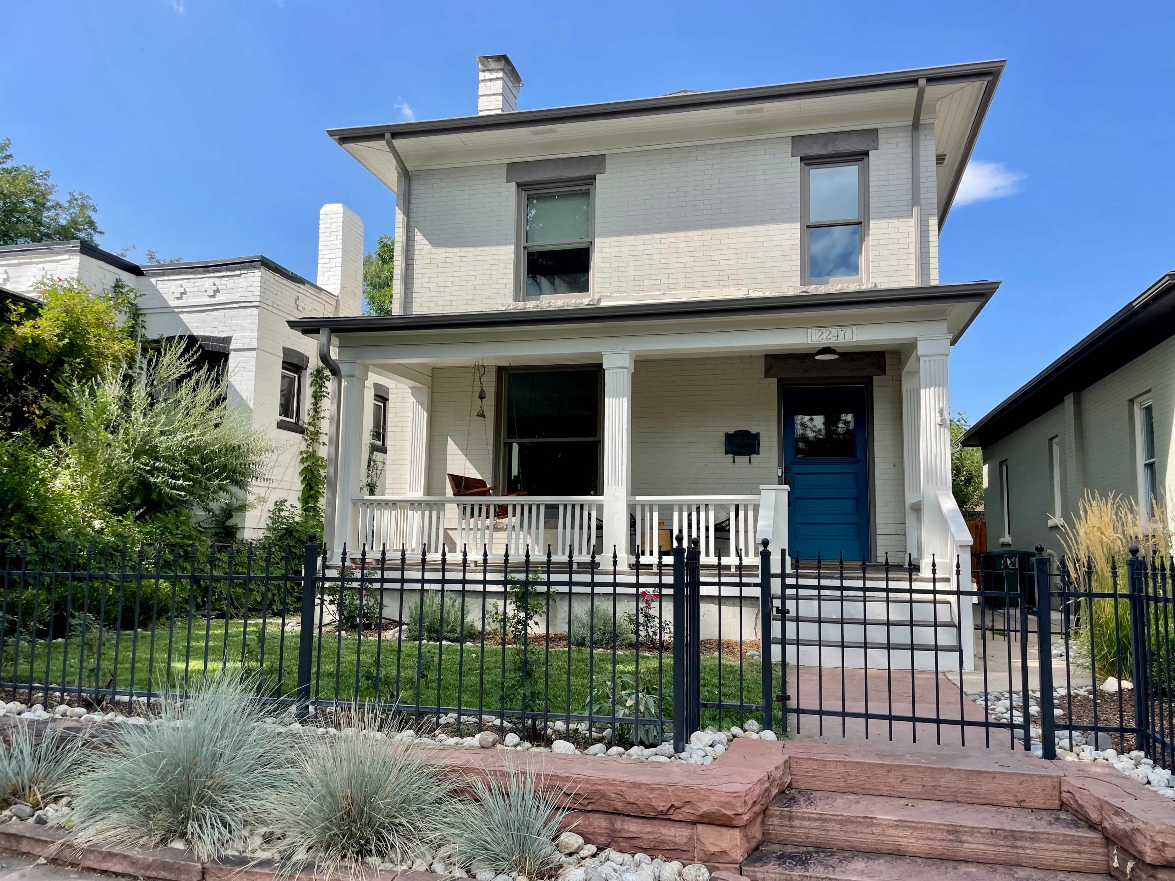 A two-story white house with a blue front door and a porch, surrounded by a manicured garden and a black wrought-iron fence.