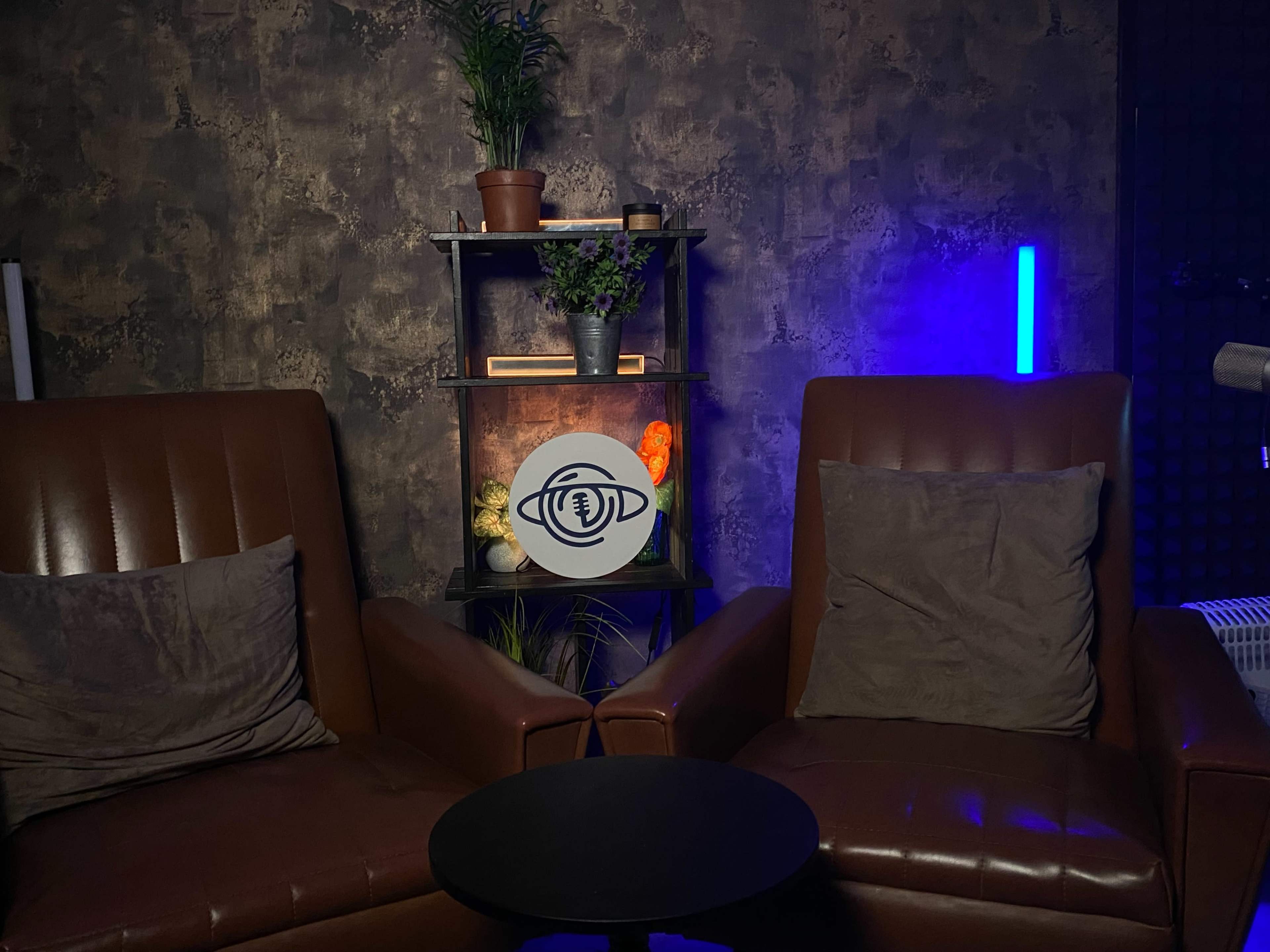 The image shows a cozy seating area with two brown leather chairs facing a small black table, flanked by shelves holding decorative plants and illuminated by colorful lights.
