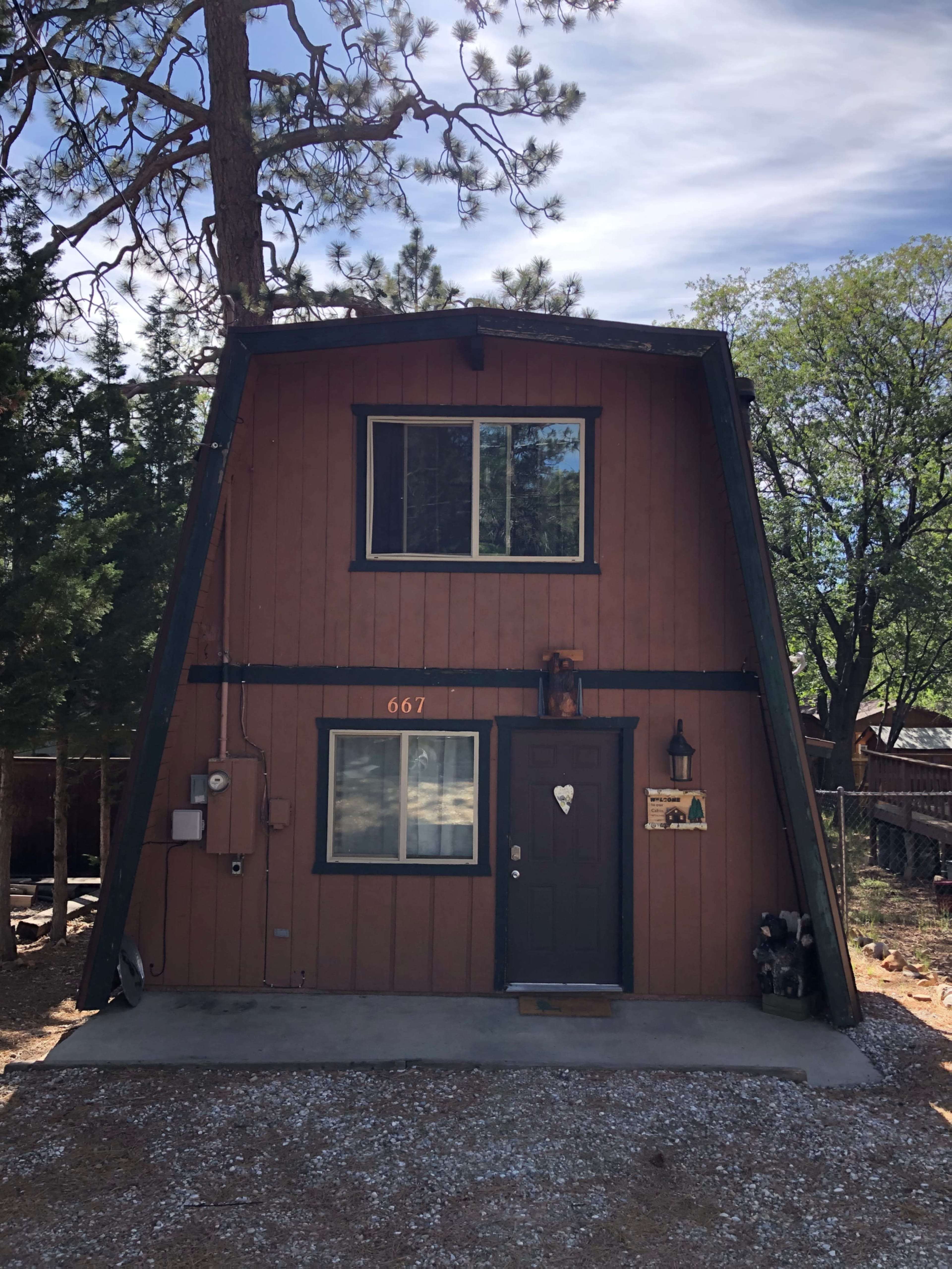 The image shows an A-frame cabin with a brown wooden exterior, featuring a front door with a heart-shaped cutout and a number "667" displayed above it.