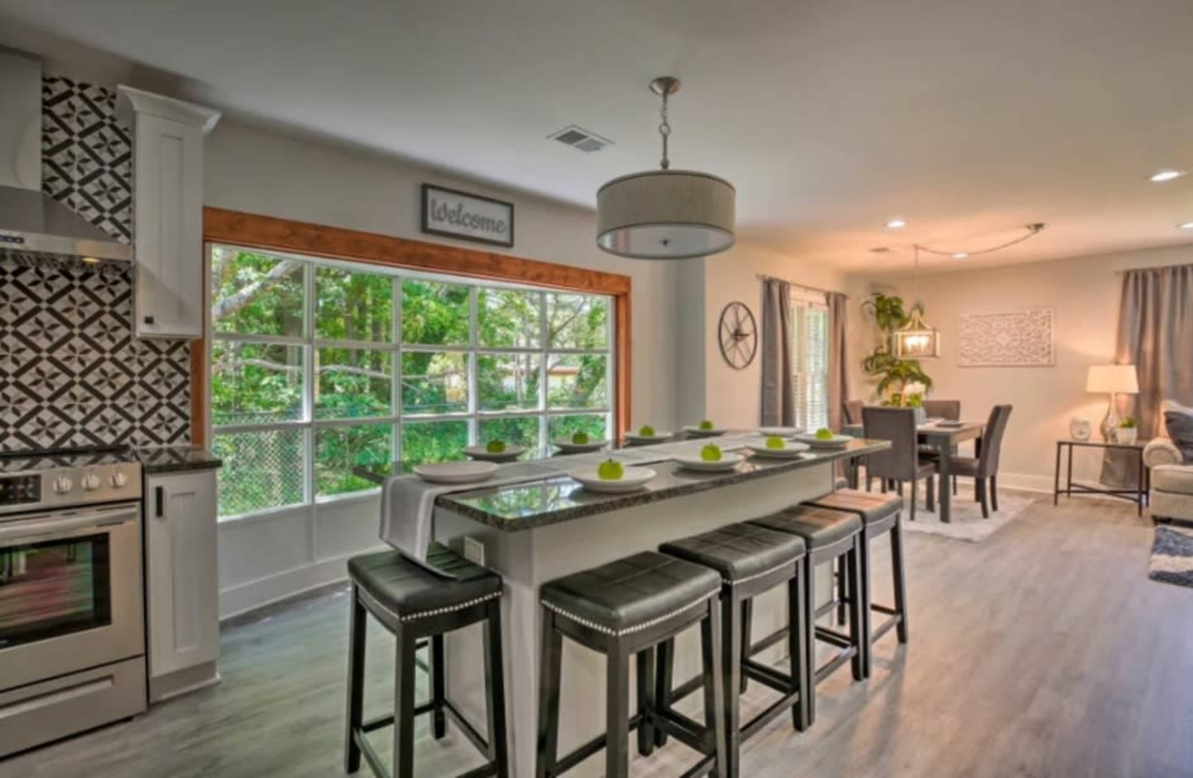 A modern kitchen with a large island, four black stools, and a living area in the background featuring a dining table and a cozy seating arrangement.