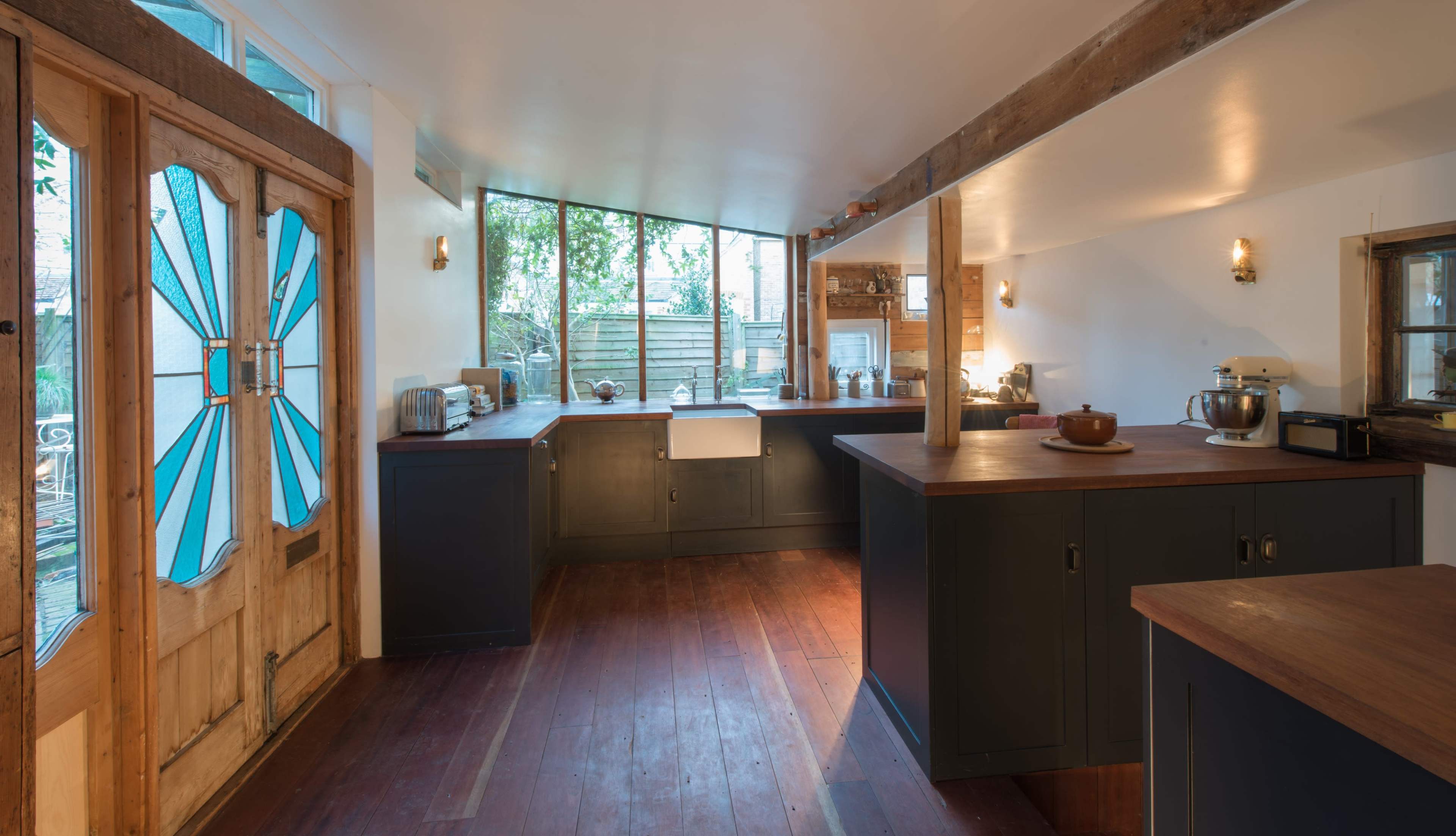 The image shows a modern kitchen with wooden flooring, dark cabinetry, and large windows that let in natural light.