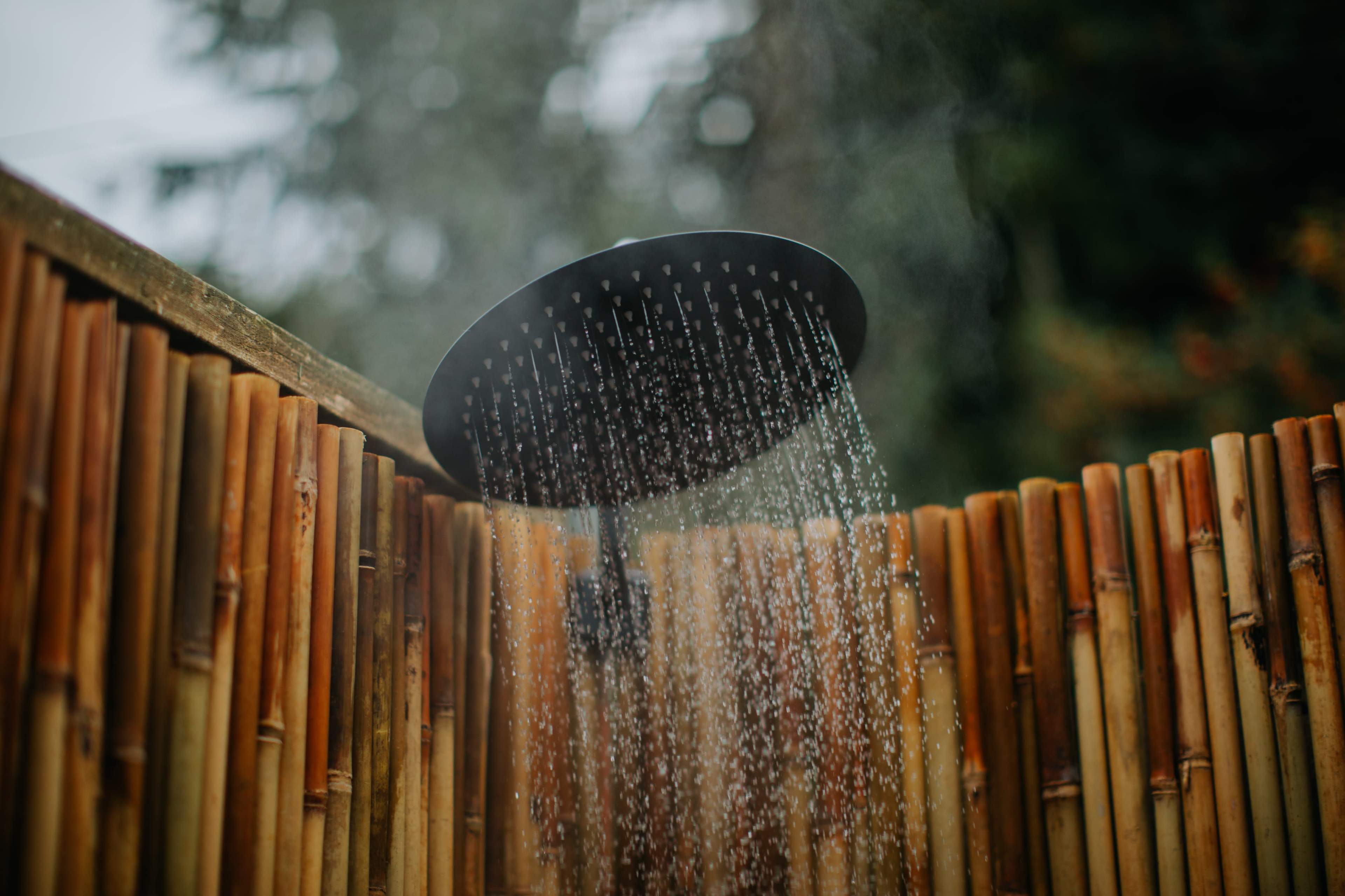 A rainfall showerhead releases water in an outdoor bamboo shower enclosure.