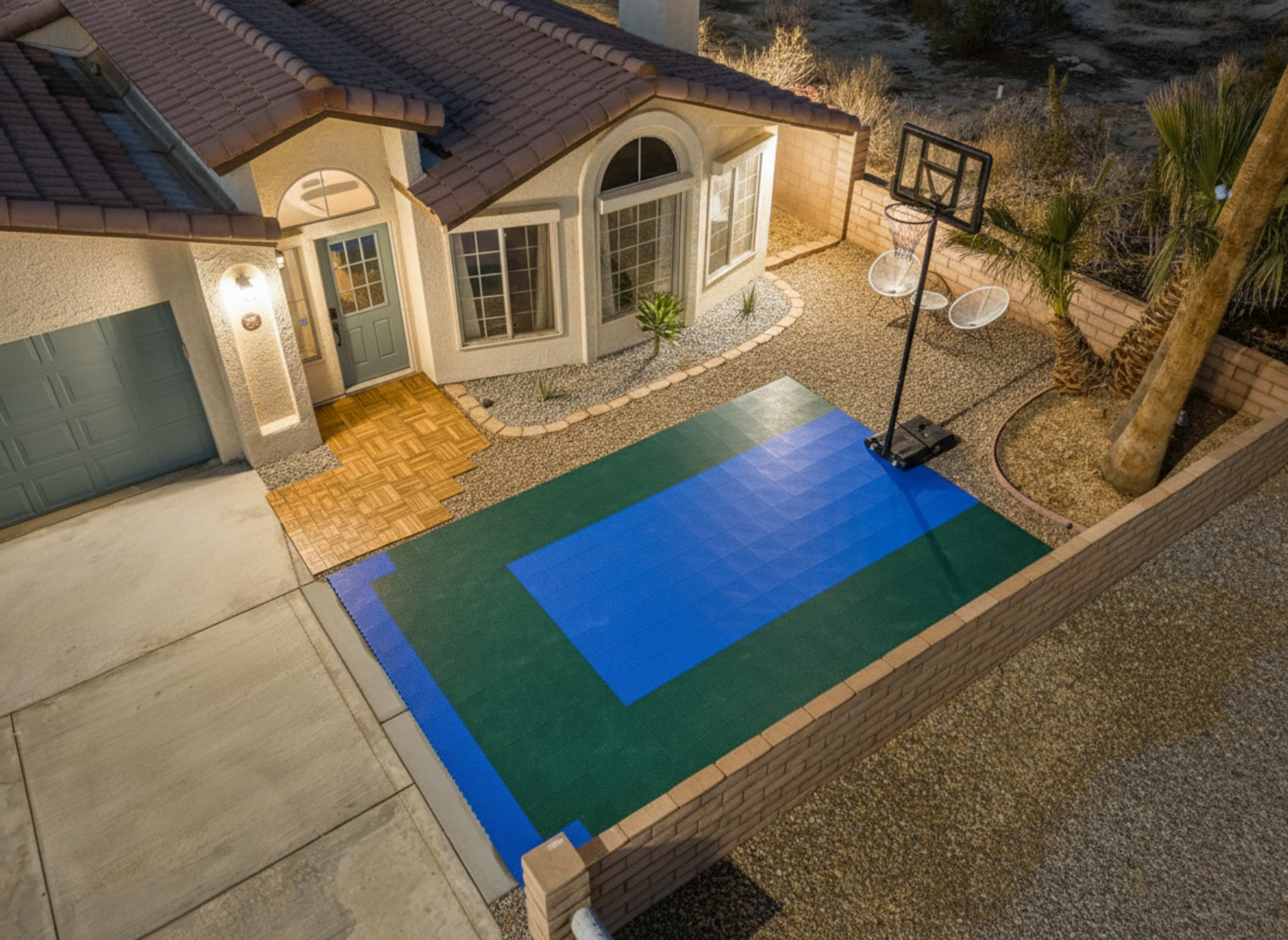 The image shows a residential exterior featuring a house with a porch and a basketball hoop set up on a green and blue court in the yard.