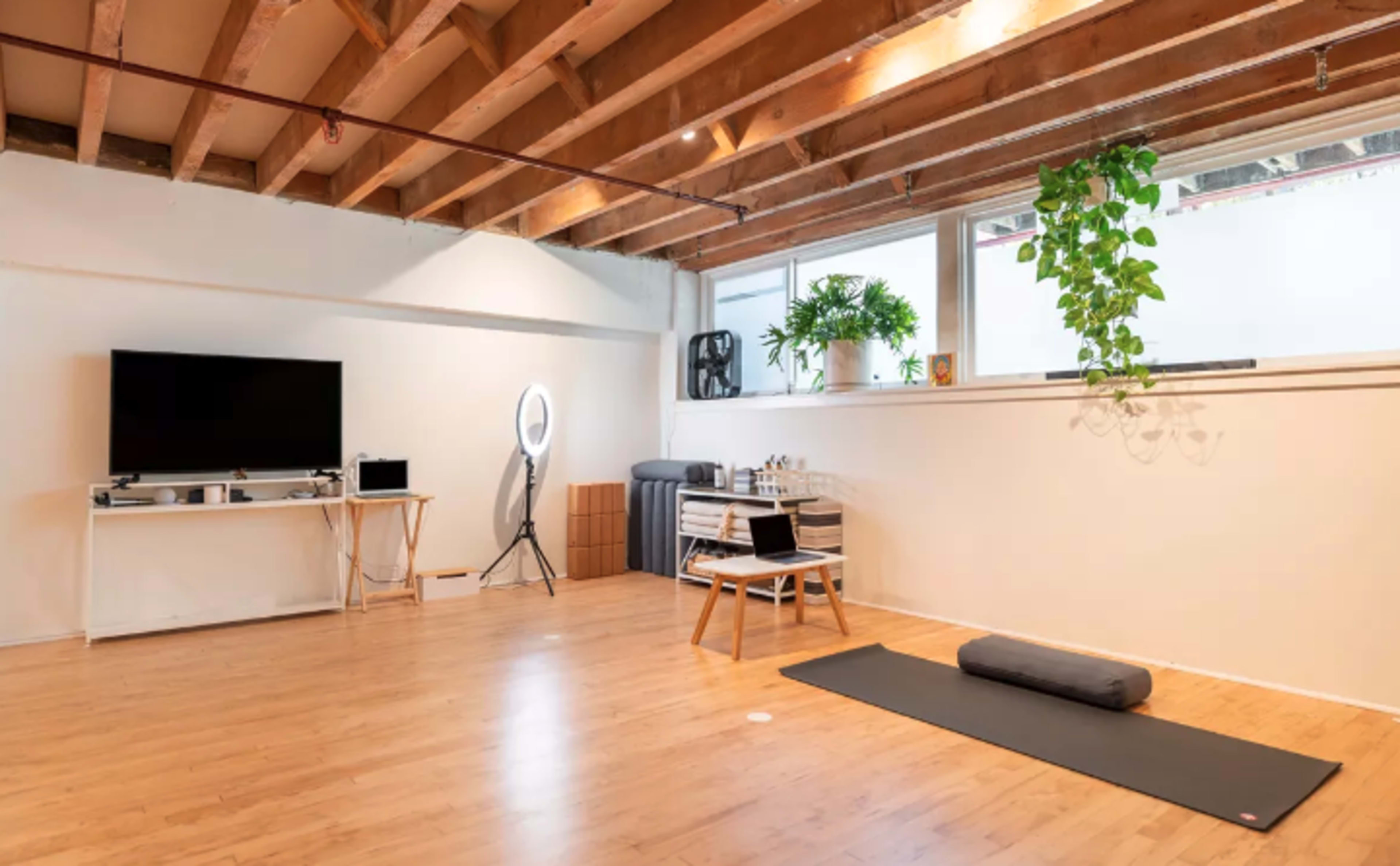A spacious, minimalist room with wooden beams, a television on a white stand, a yoga mat, and a large window with plants.
