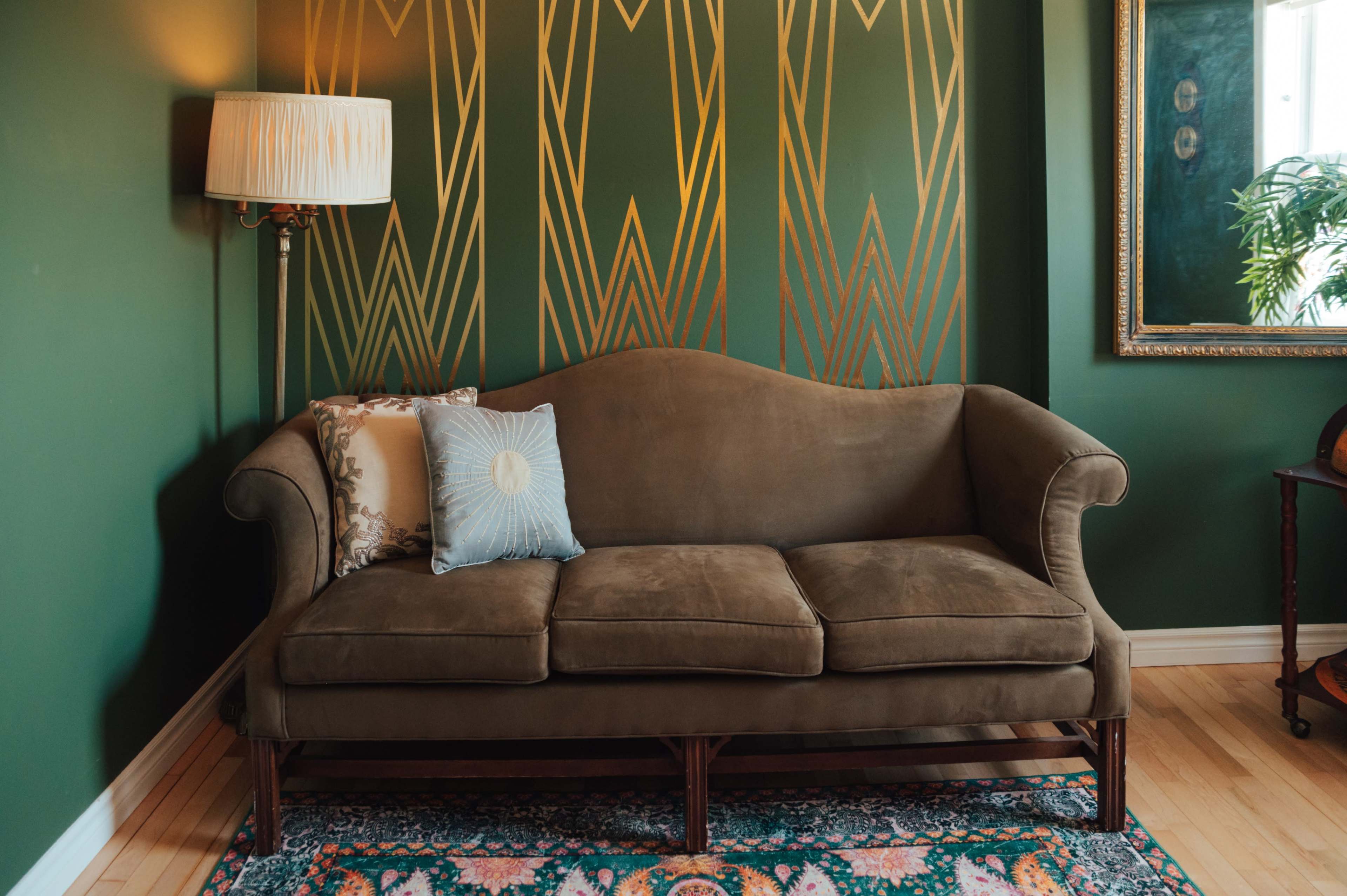 A brown sofa with a decorative pillow sits against a green wall featuring gold geometric designs, complemented by a lamp and a framed mirror in the room.