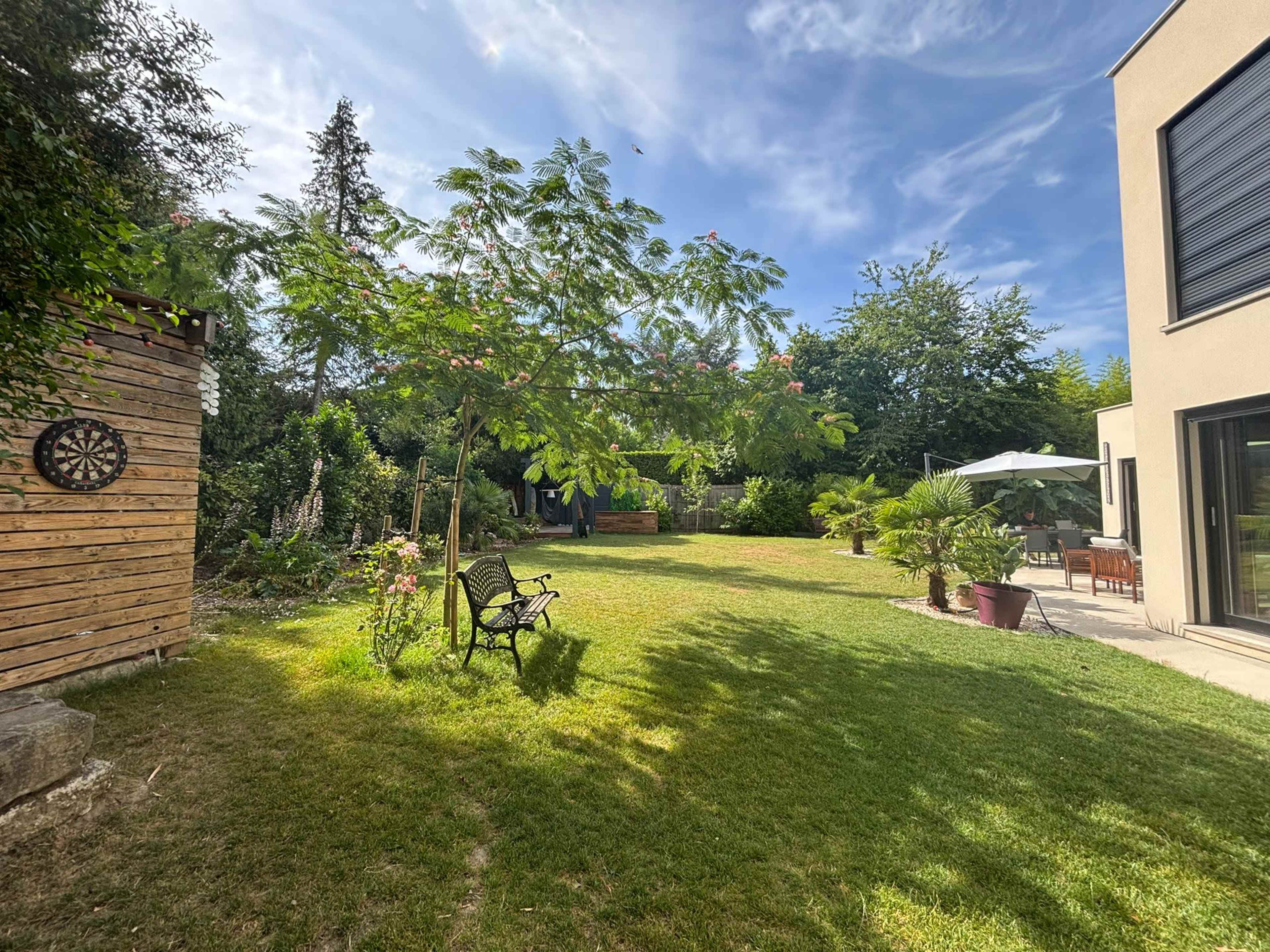 The image shows a well-maintained garden with a grassy area, a wooden board with a dartboard, a bench, and a patio with outdoor furniture and an umbrella.
