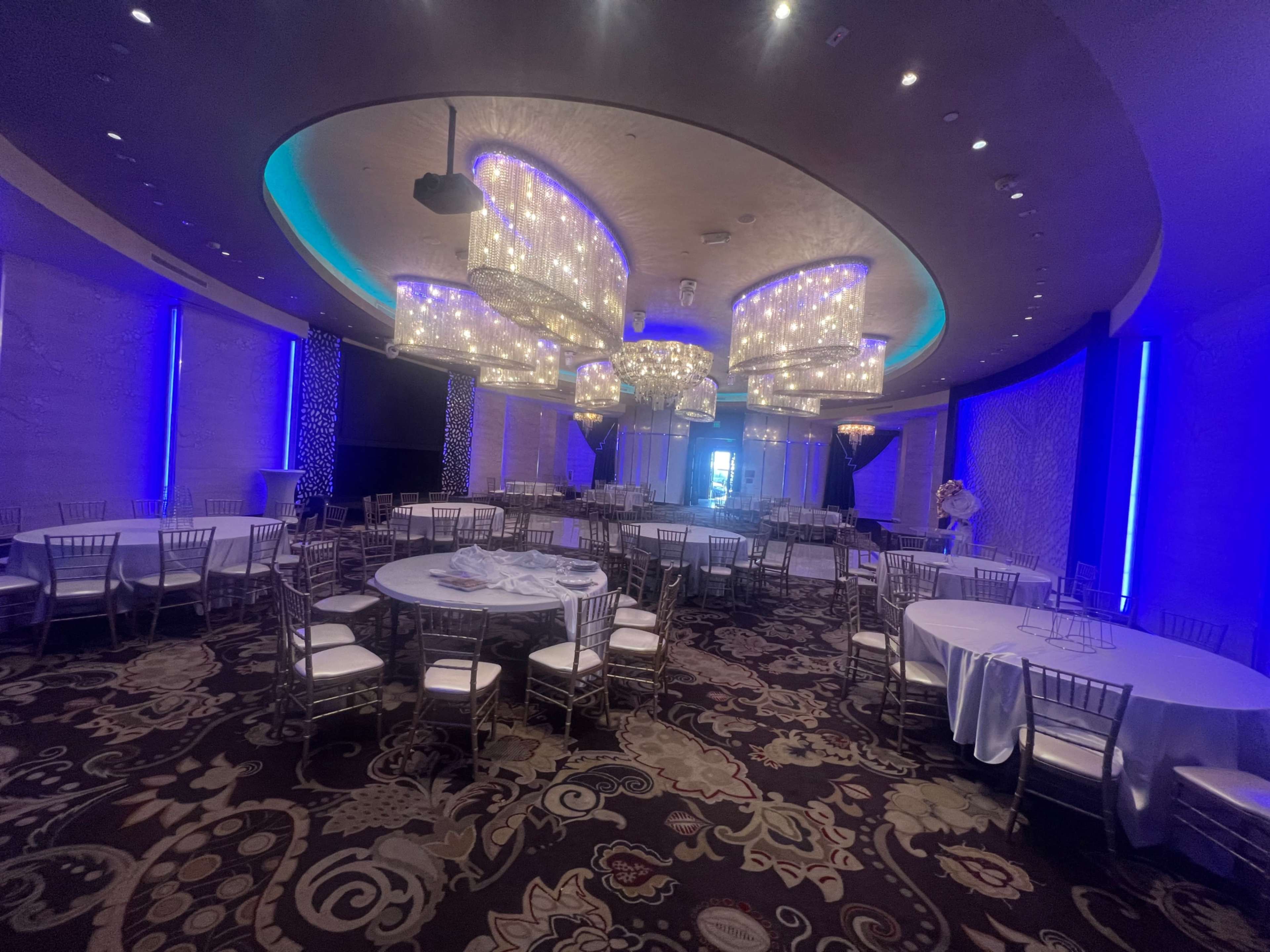 An elegantly arranged event space with several round tables and sparkling chandeliers under blue lighting.