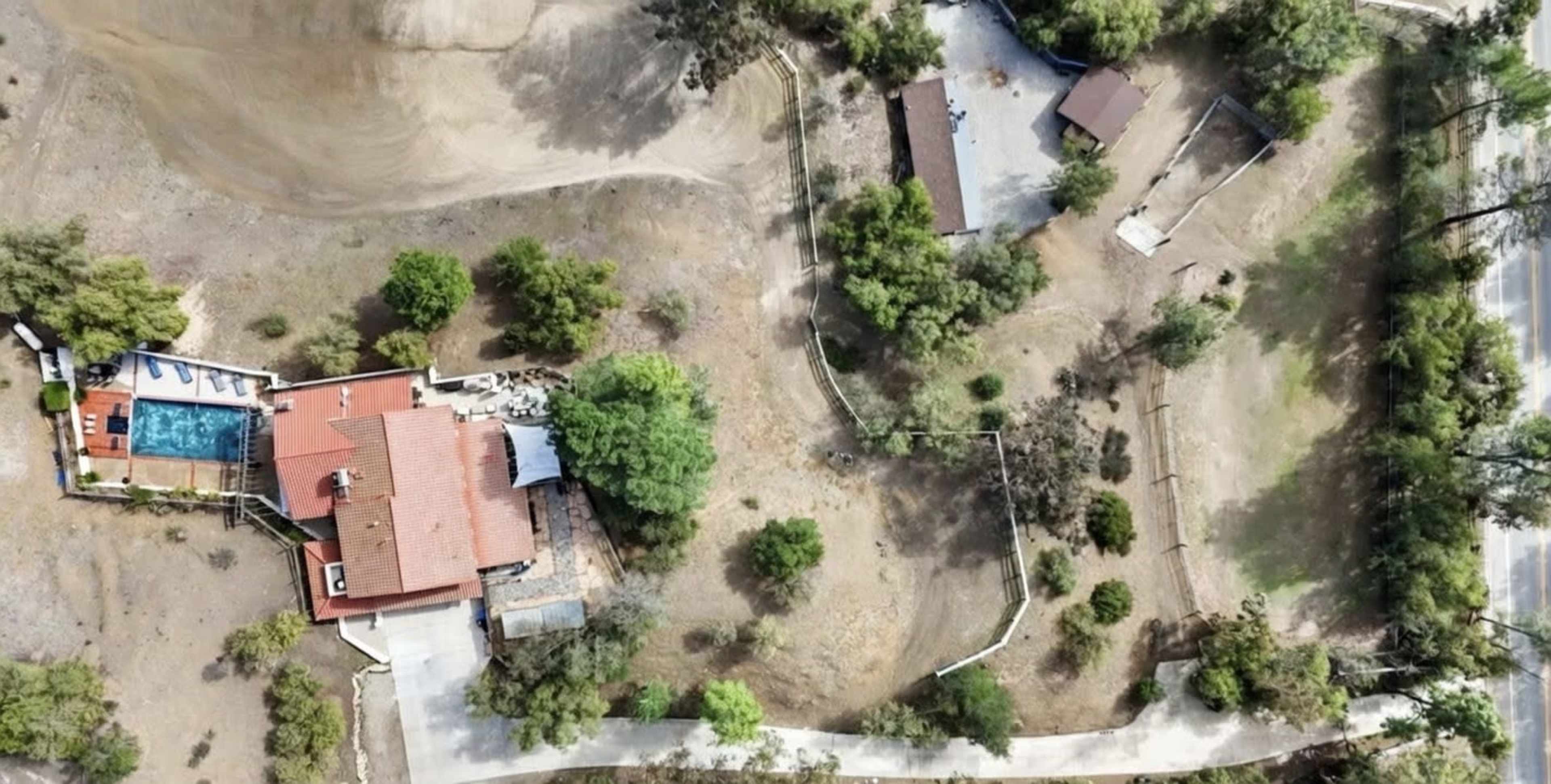 The aerial view shows a large property with a main house, a swimming pool, and a smaller nearby building surrounded by sparse landscaping and bare ground.