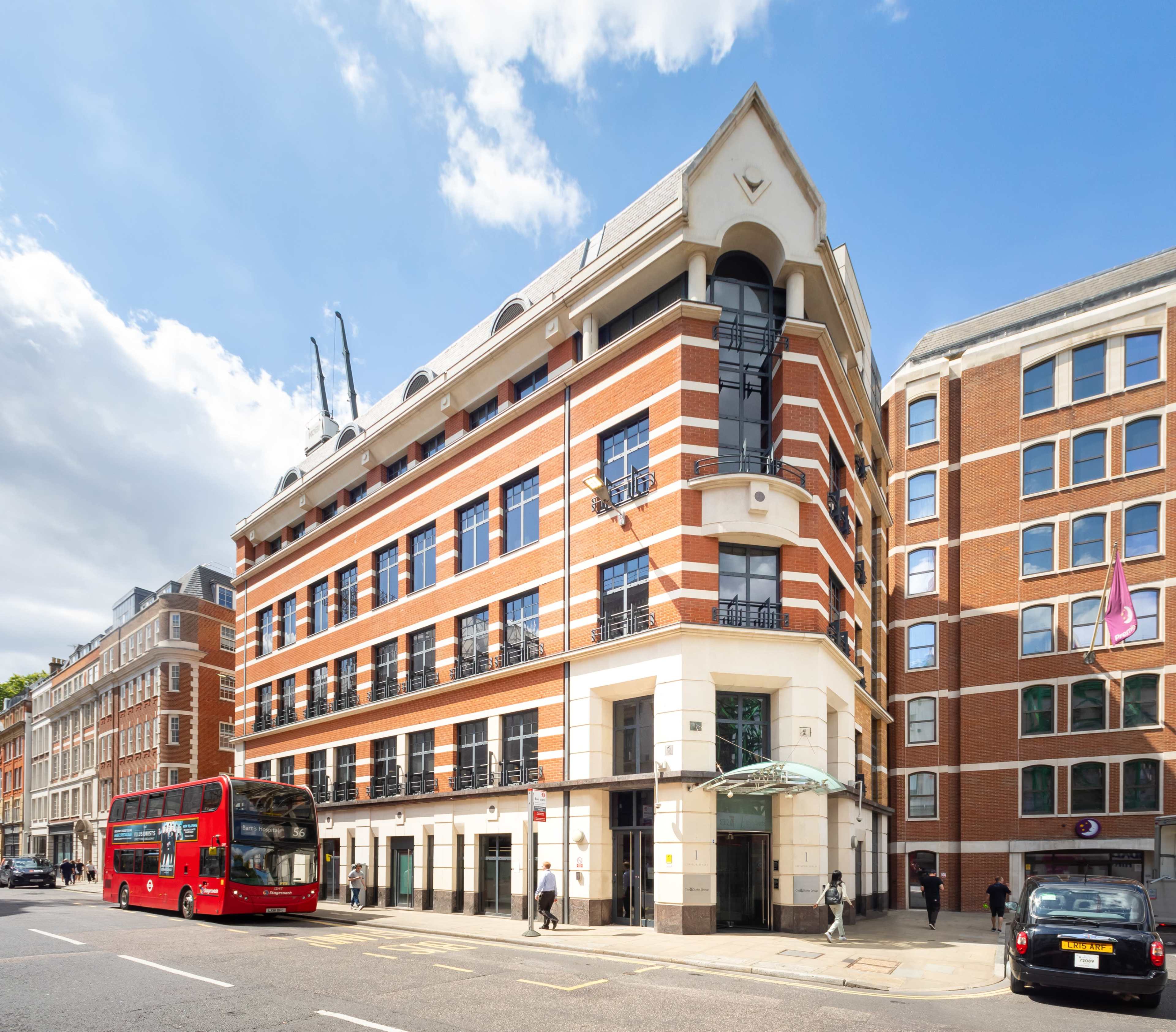 A red double-decker bus stops in front of a modern brick building with balconies and shops on the ground floor, set against a partly cloudy sky.