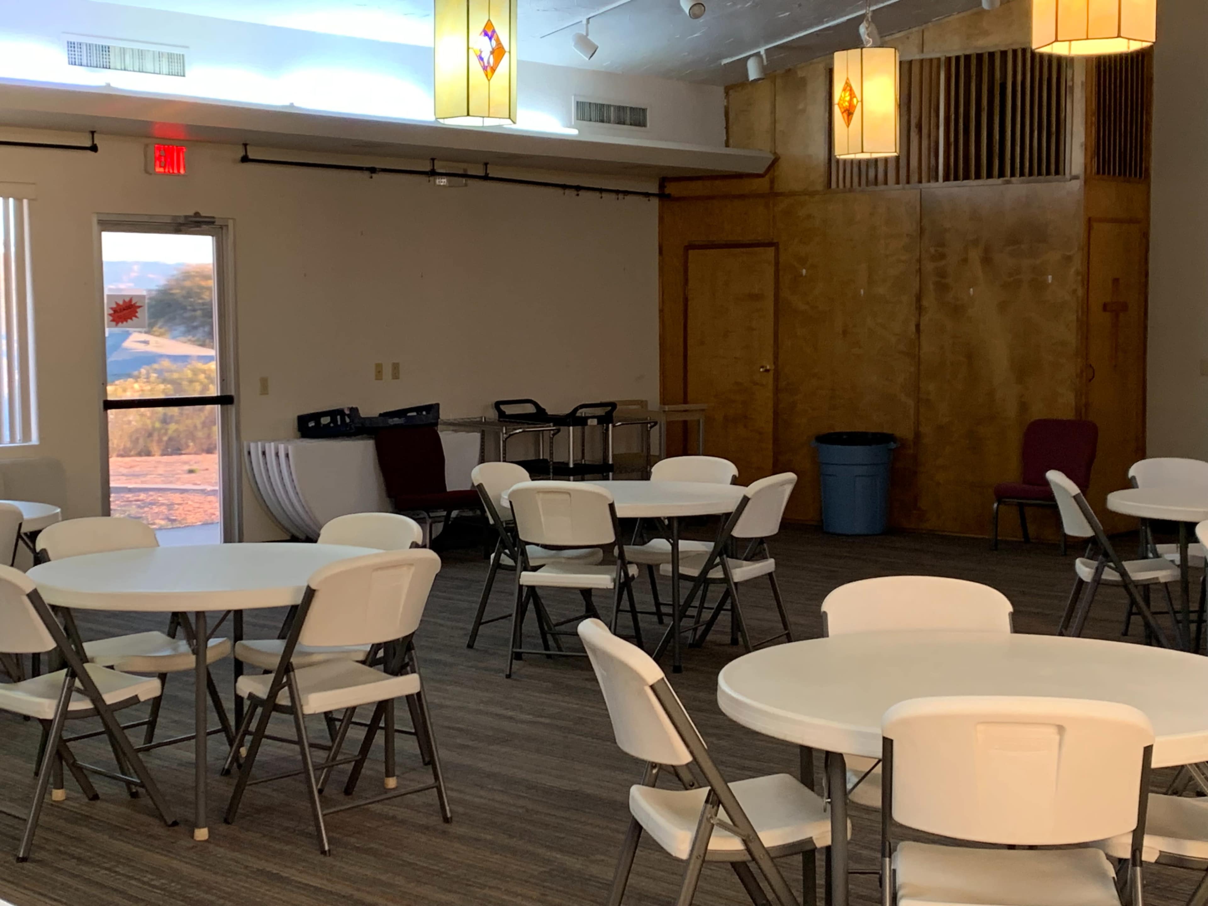 The image shows a large indoor room with round tables and folding chairs arranged, including a doorway leading outside and a blue trash can in the corner.