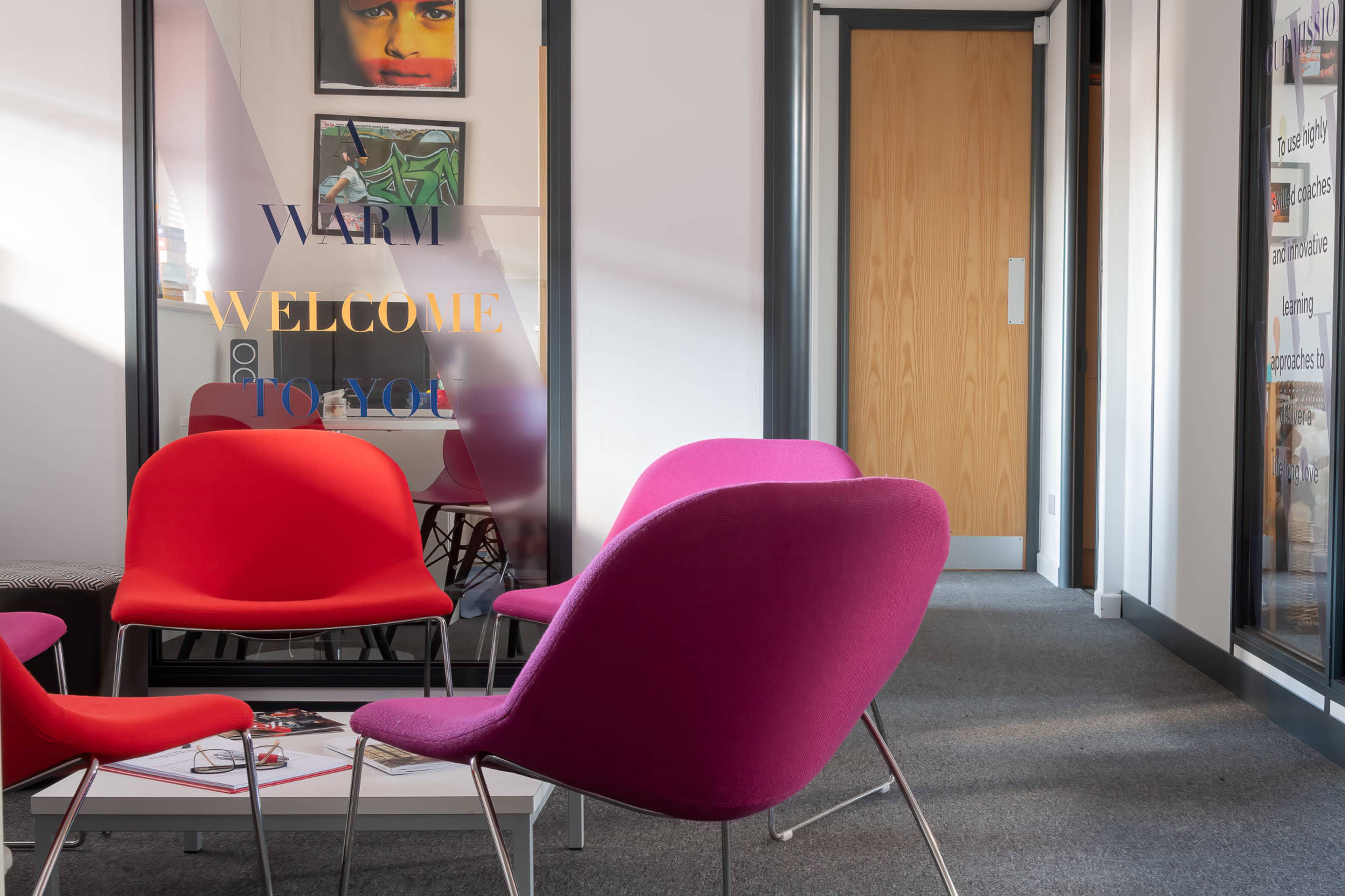 The scene features colorful chairs in a waiting area with a glass partition displaying the words "WARM WELCOME TO YOU."