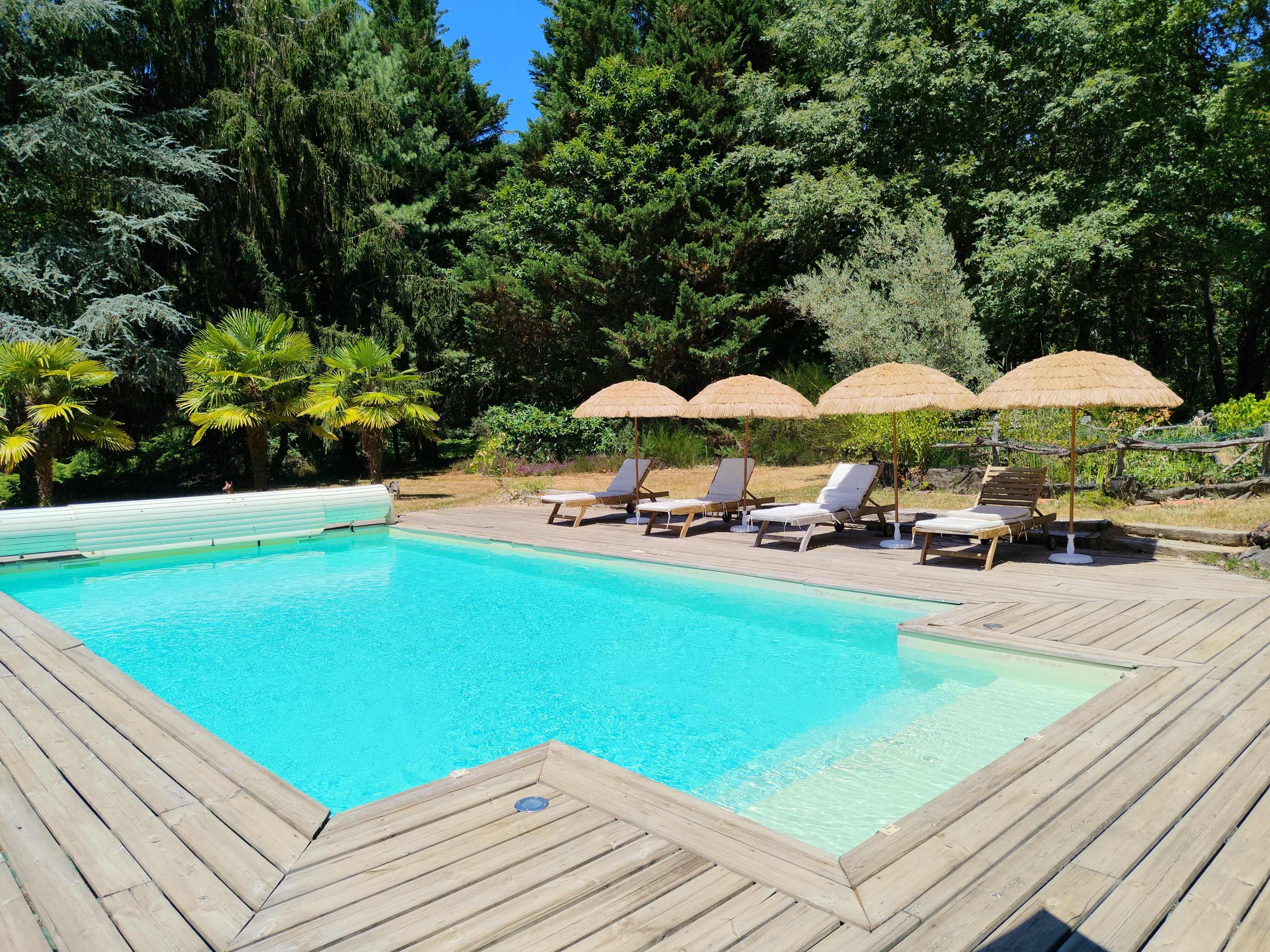 A swimming pool with wooden decking is surrounded by lounge chairs and parasols in a shaded outdoor setting.
