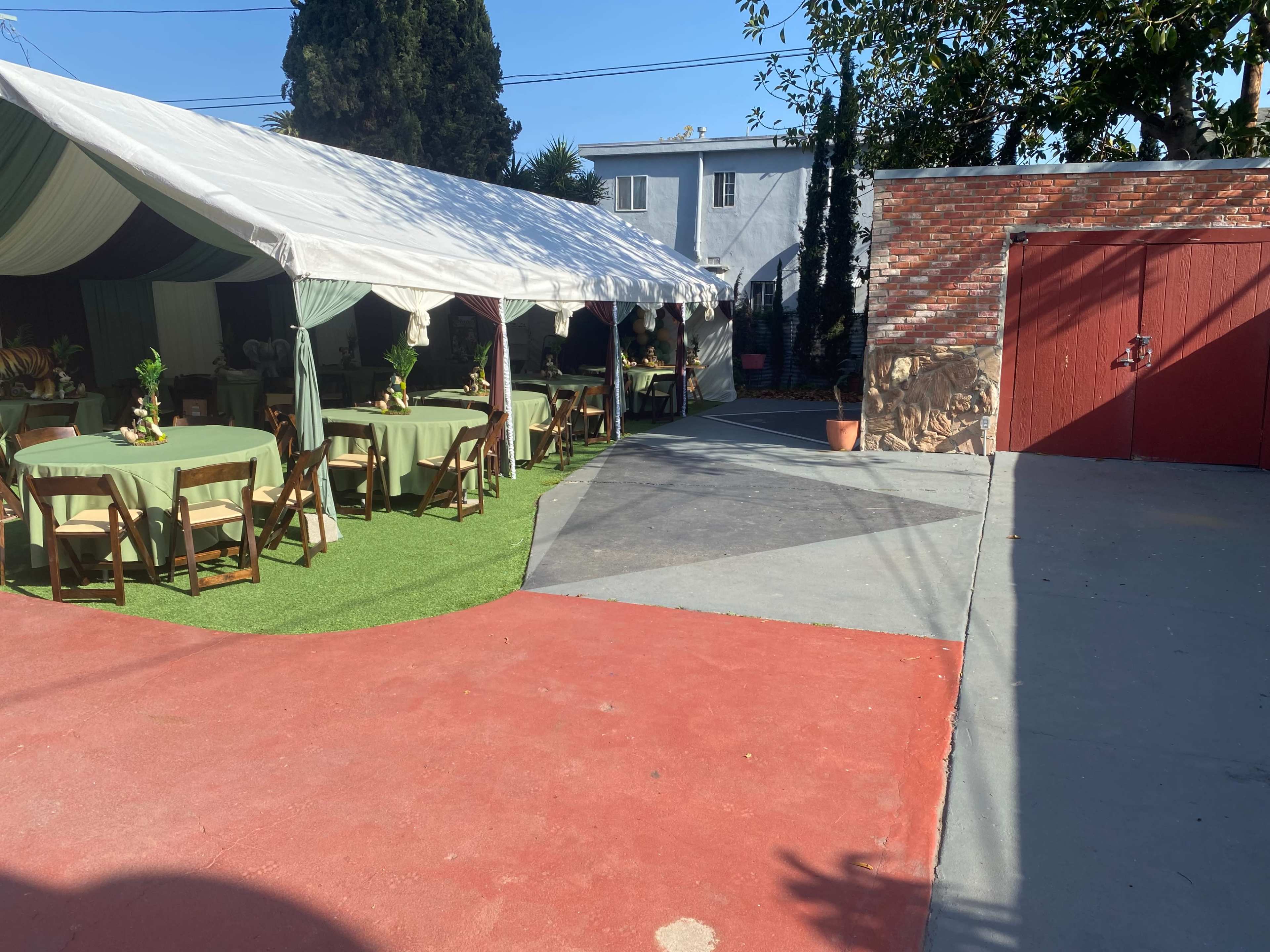 A large outdoor tent with tables set up under it, alongside a paved area leading to a red garage door.