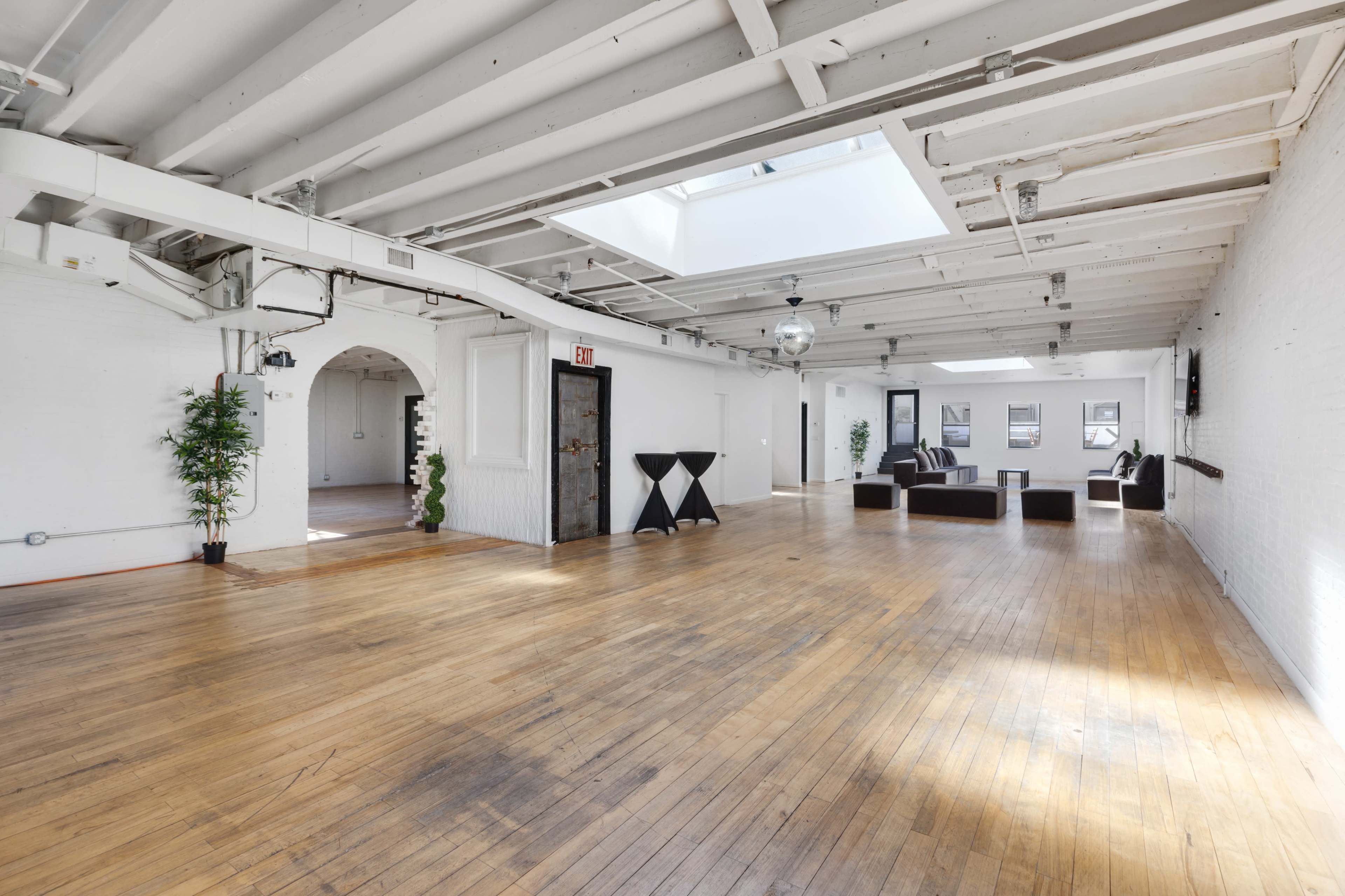 A spacious, airy interior with wooden floors, white walls, a skylight, and minimal furniture, including a black table and a seating area.