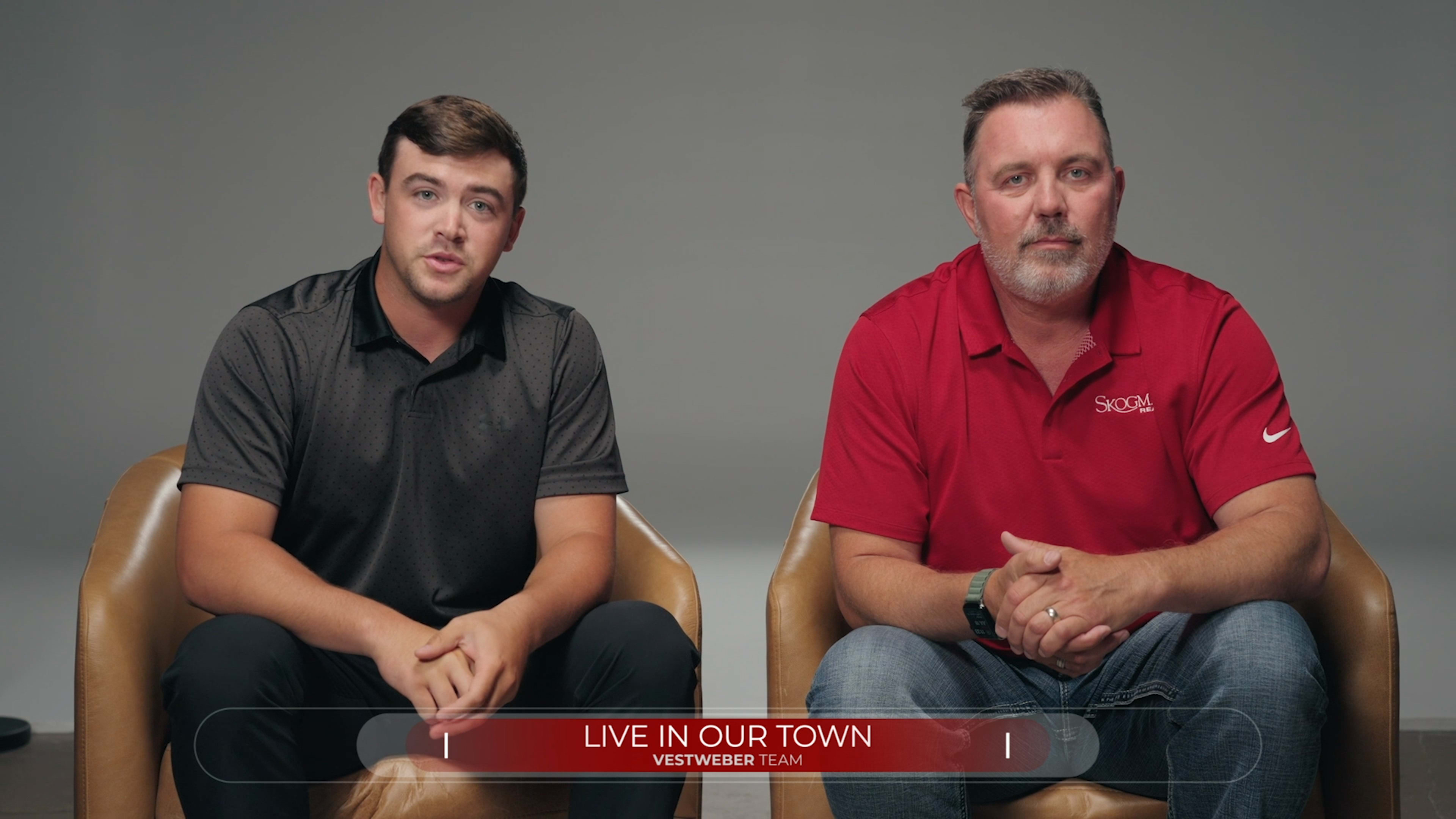Two men sit side by side on brown chairs against a gray backdrop, the one on the left wearing a black shirt and the one on the right in a red polo, both looking at the camera.