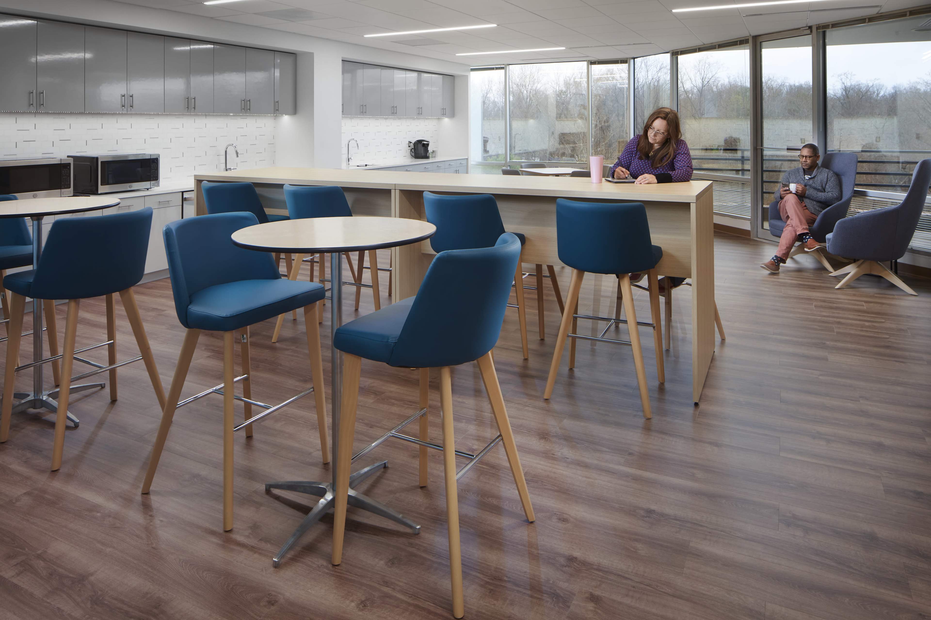 A modern break room features high tables and chairs, with one person working on a laptop and another seated nearby holding a cup.