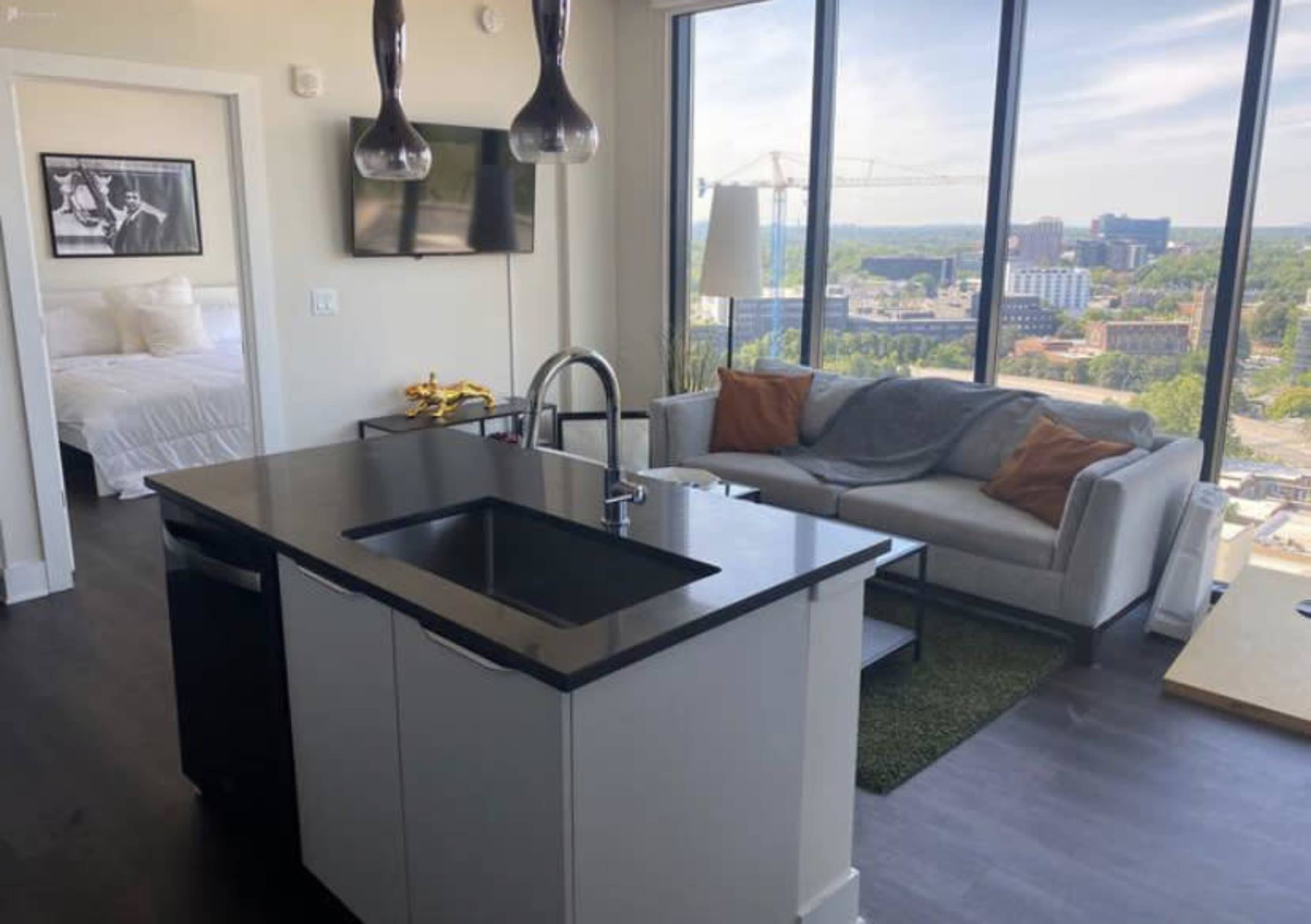 The image shows a modern apartment interior with a kitchen island, a living area featuring a sofa and a television, and a bedroom visible in the background through large windows.